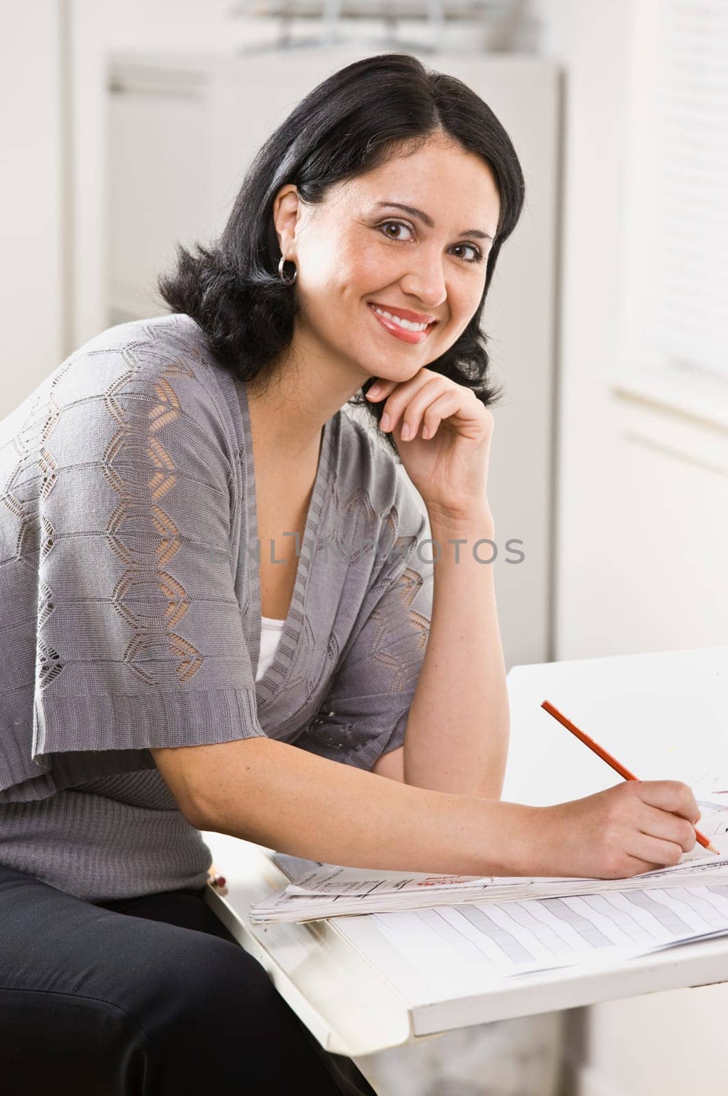 A young, attractive woman is looking over some paperwork on a desk, and smiling at the camera.  Vertically framed shot.