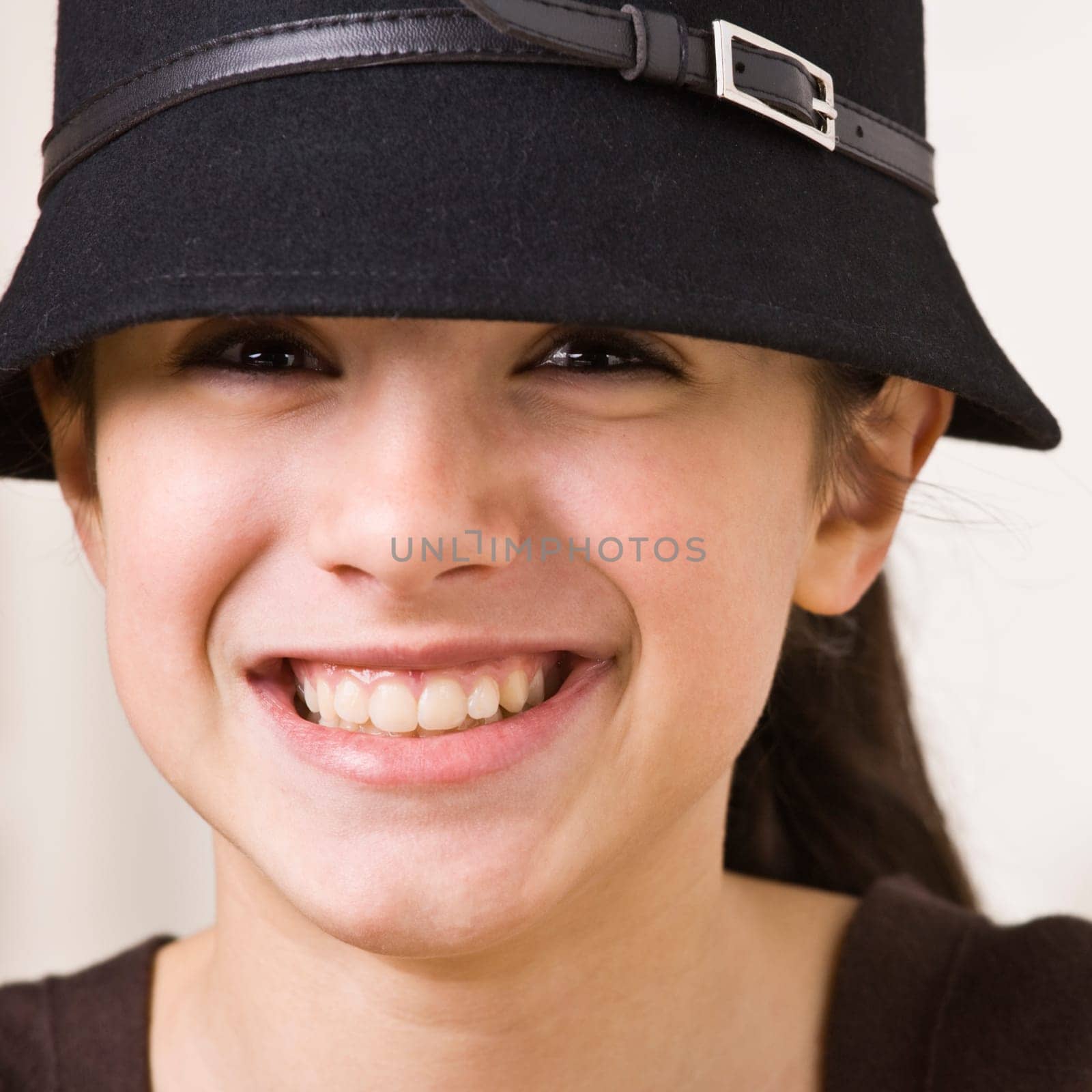 A young girl is wearing a hat and smiling at the camera.  Square composition.