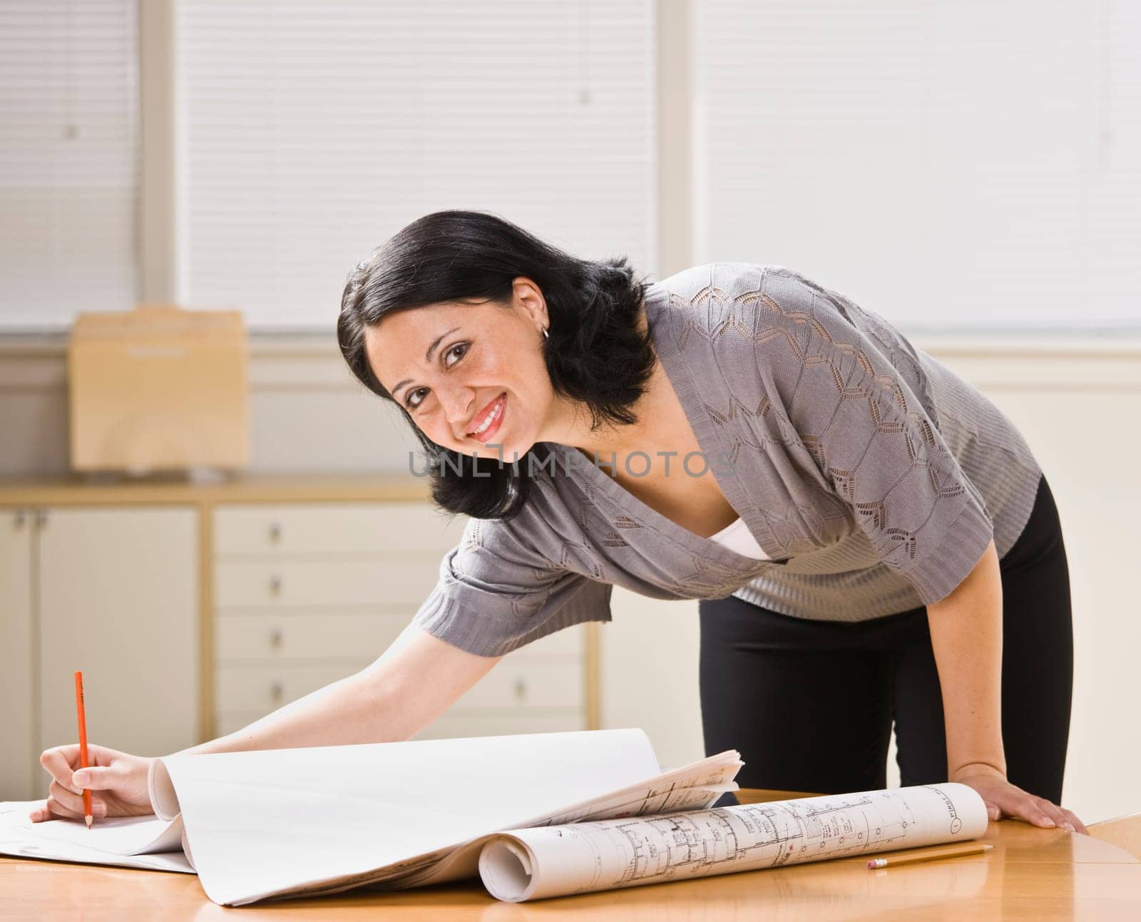 Attractive woman leaning over desk with drawings. Pen in hand, looking at camera. Horizontal.
