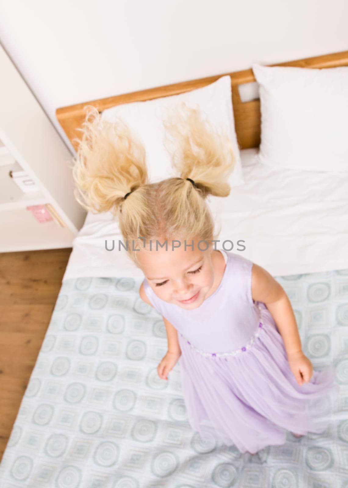 A young girl is jumping up and down on a bed.  She is smiling and looking away from the camera.  Vertically framed shot.