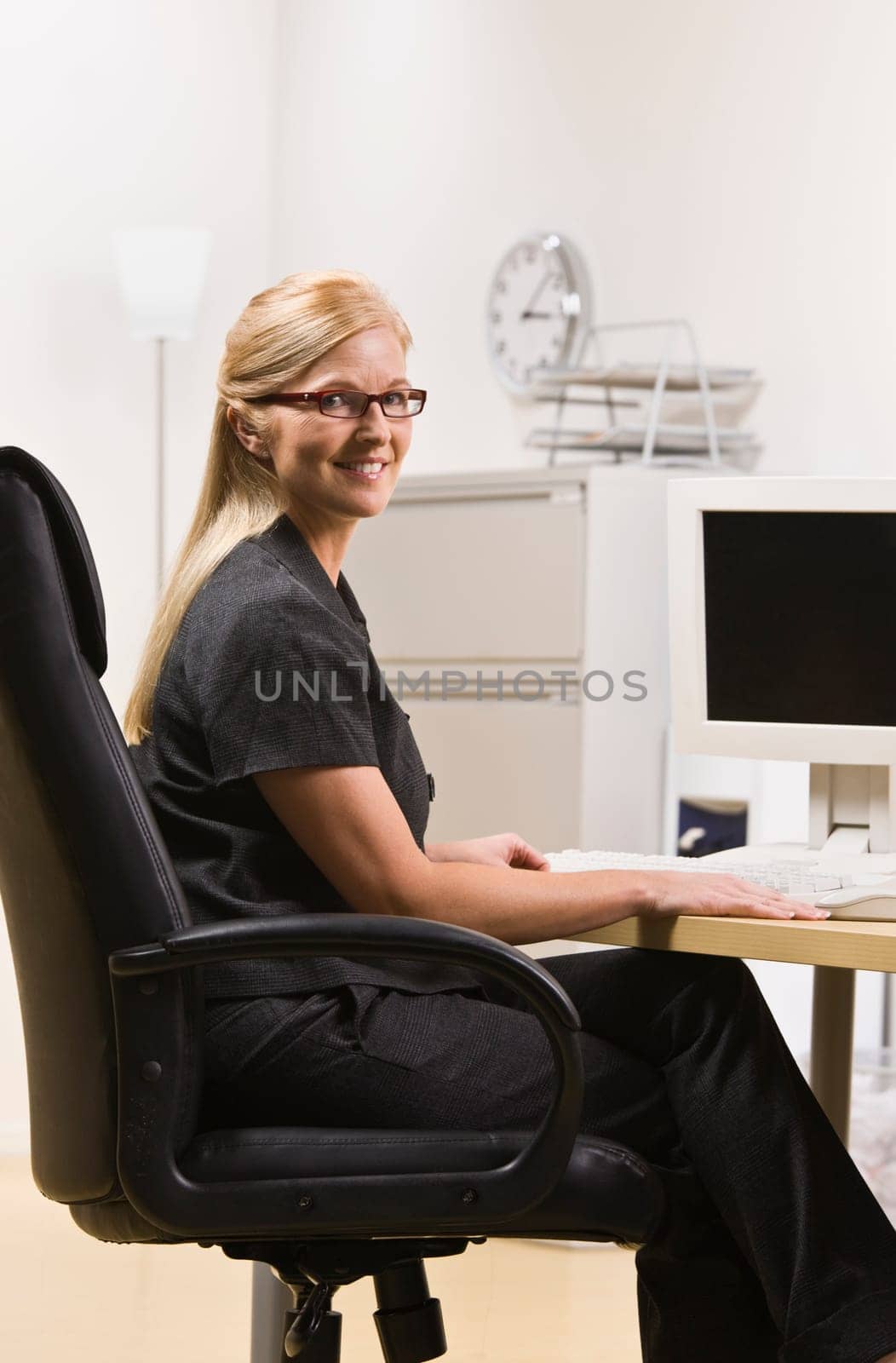 A businesswoman is seated at a computer desk and smiling at the camera.  Vertically framed shot.