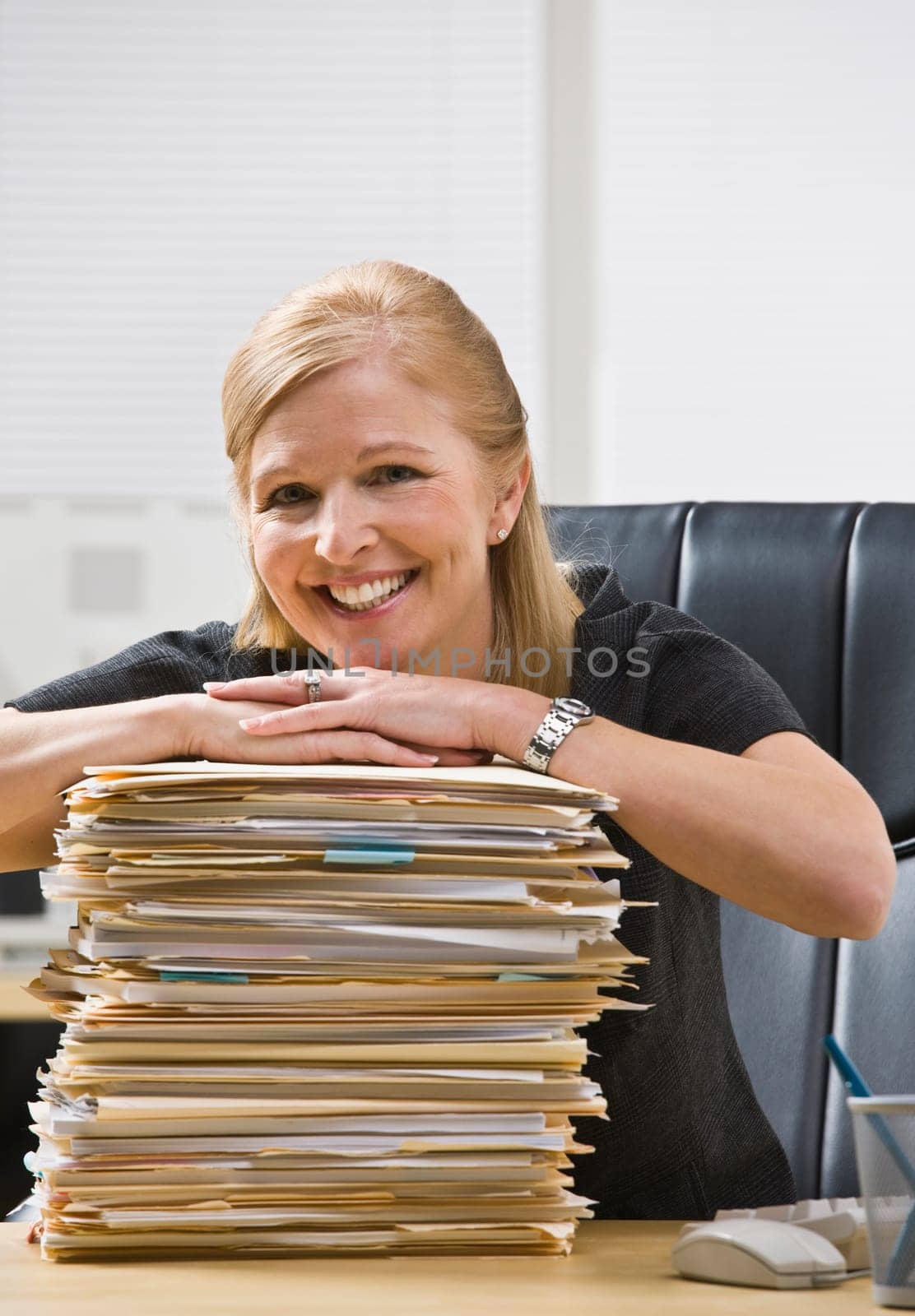 A businesswoman is seated at a desk in an office with a stack of paperwork.  She is smiling at the camera.  Vertically
