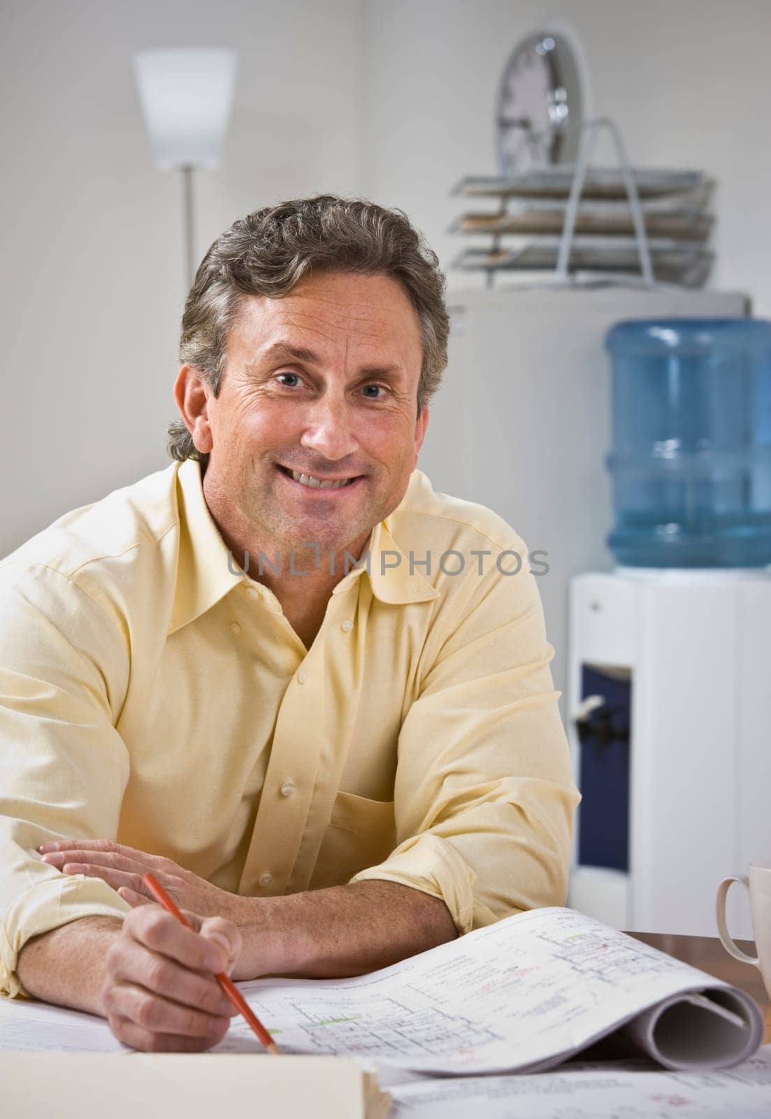 A man is sitting at a desk and working on blueprints.  He is smiling at the camera.  Vertically framed shot.