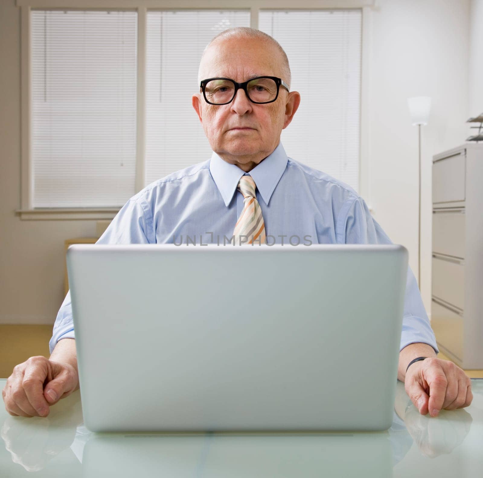 An elderly businessman is working on a laptop in an office.  He is looking at the camera.  Square framed shot.