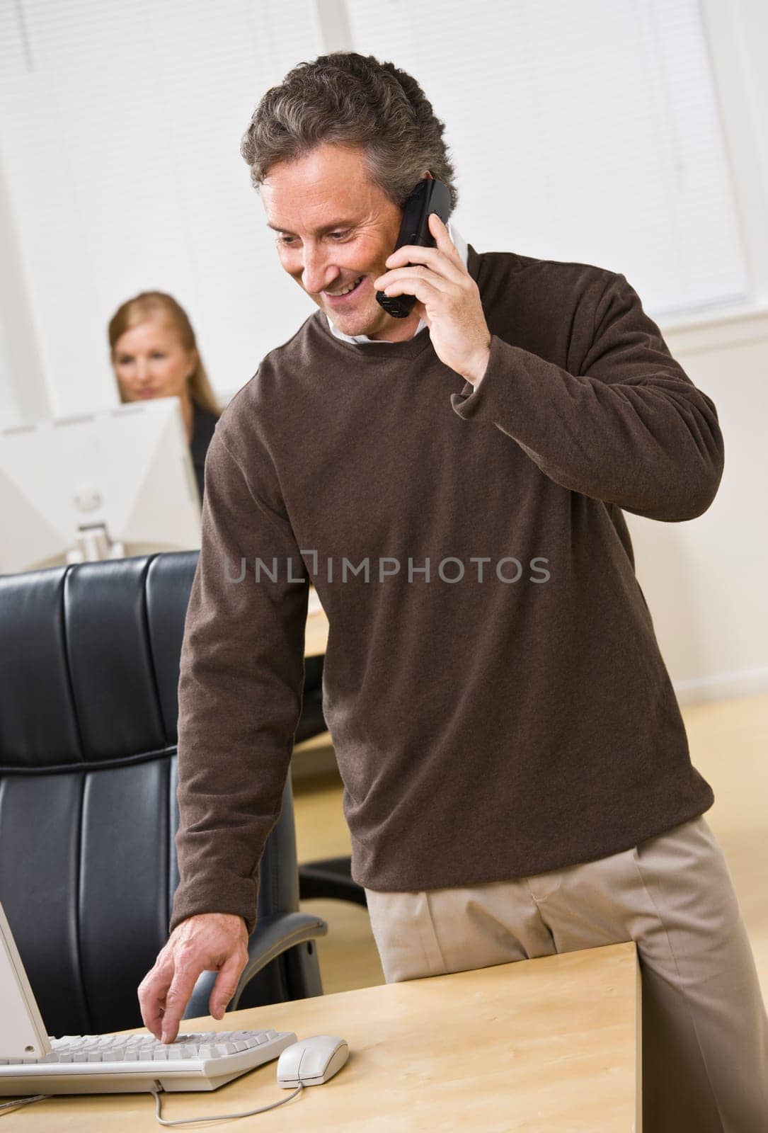 Attractive business man standing over keyboard while chatting on the phone. Woman peering in back. Looking at the computer screen. Vertical.