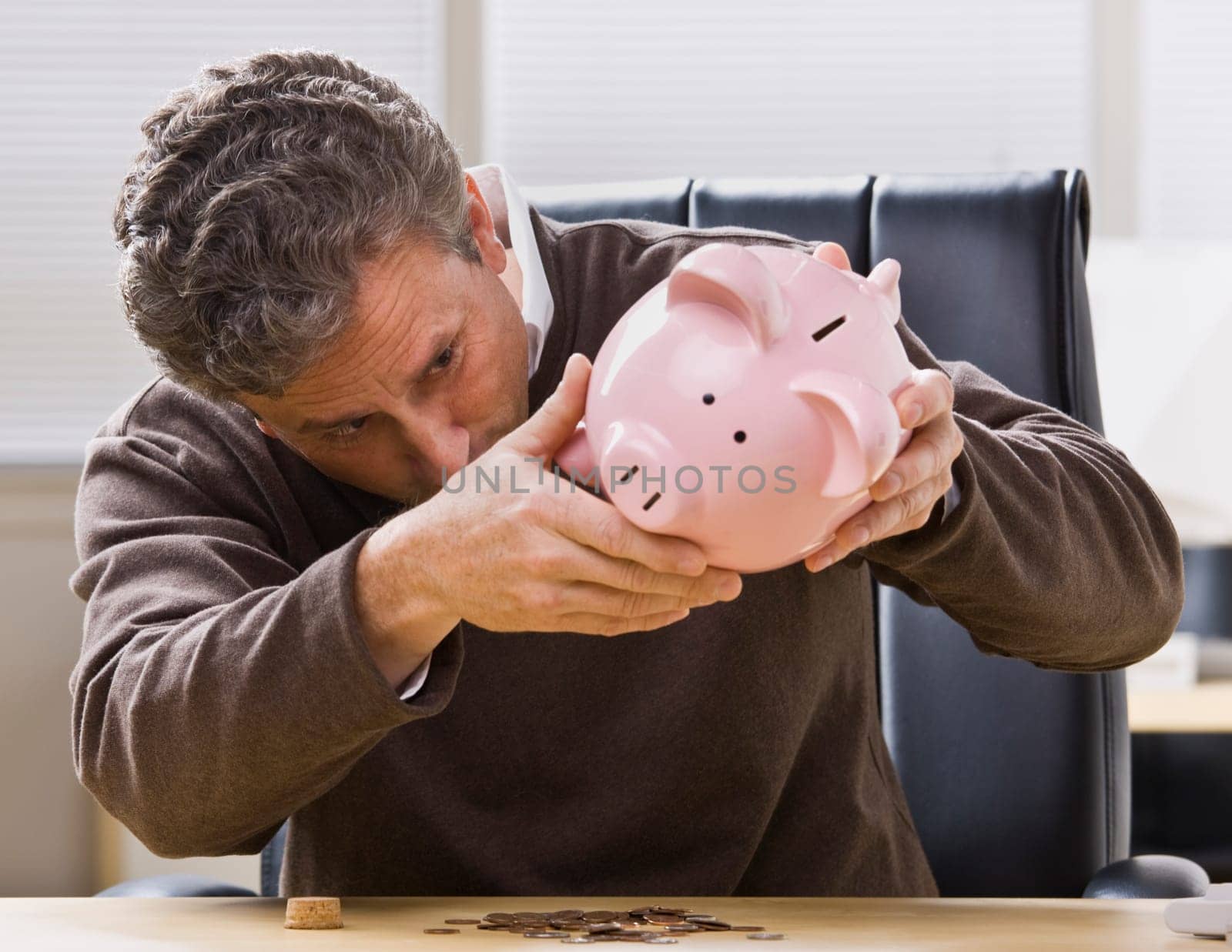 A businessman is seated at a desk in an office and is looking at a piggy bank.  Horizontally framed shot.