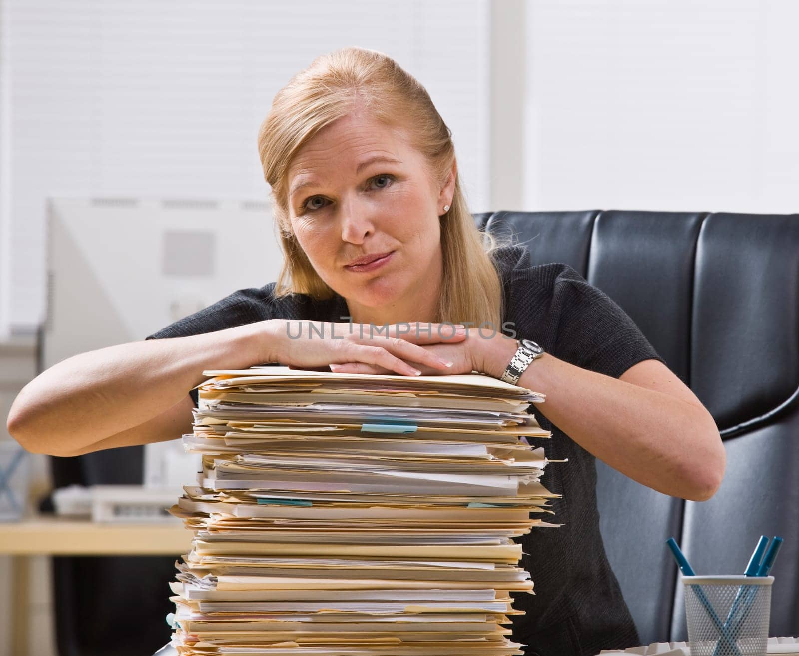 A businesswoman is seated at a desk with a large stack of paperwork in front of her.  She is looking at the camera.  Horizontally framed shot.