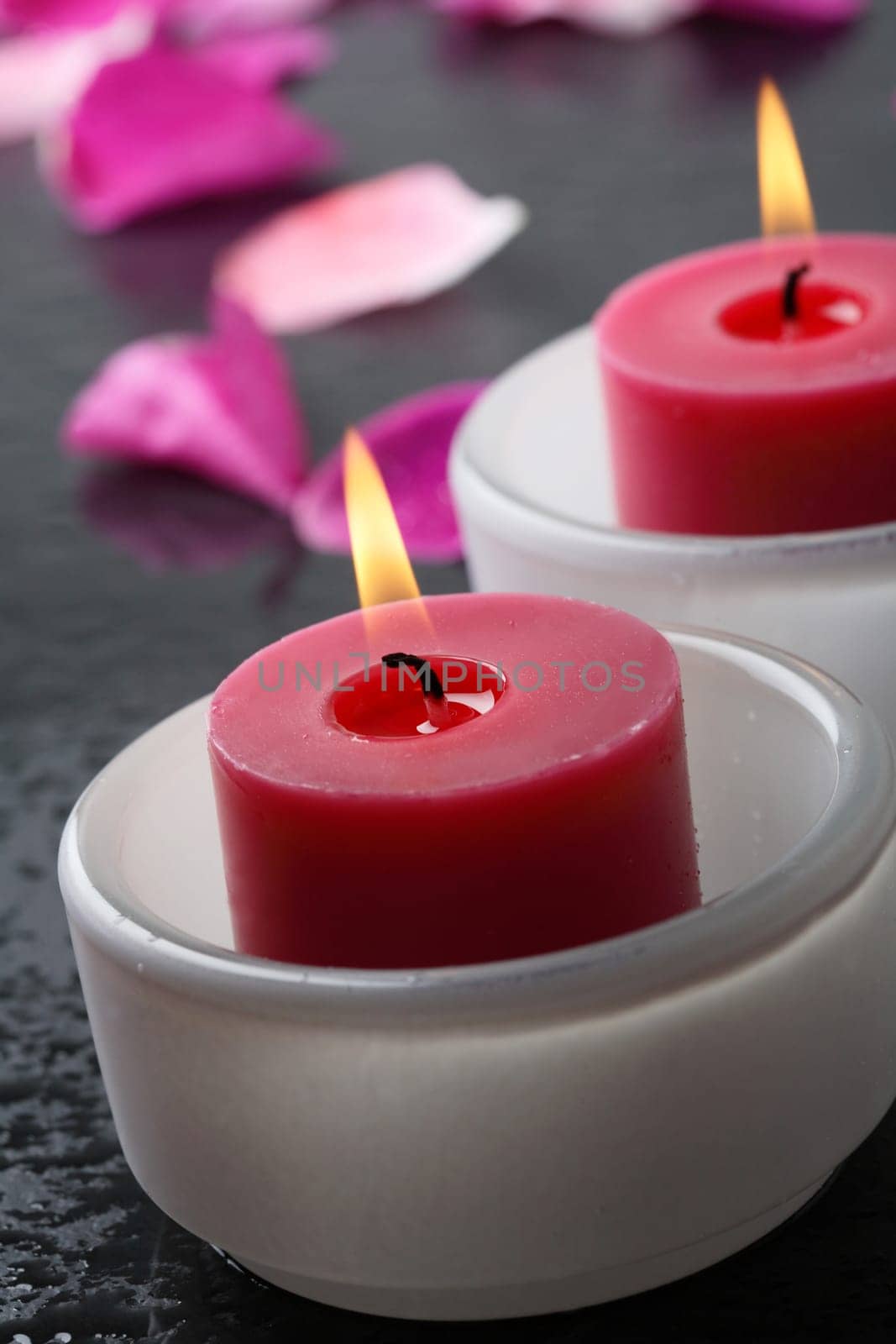 A close-up of a health and beauty spa decoration with two red colored candles in white opaque bowls surrounded by pink flower petals on a black stone surface.