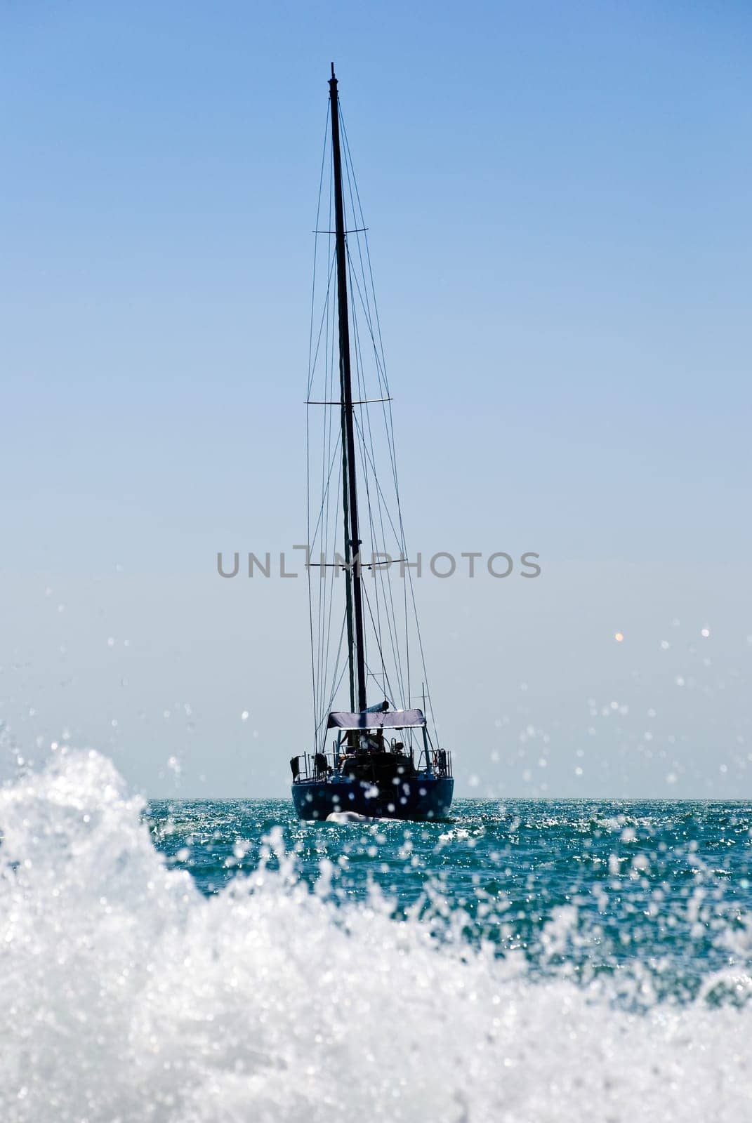 yacht drifting in the sea with sails down. Splash of water wave in foreground and blue sky in background