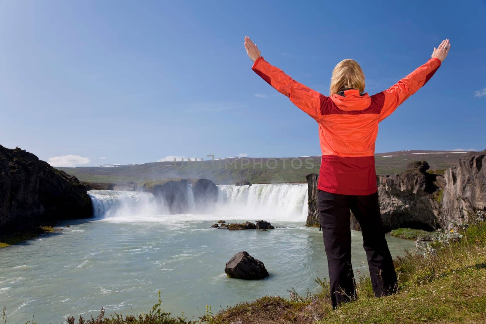 A young woman with her arms up in celebration of achievement by a waterfall. Shot on location at Godafoss waterfall in Iceland.