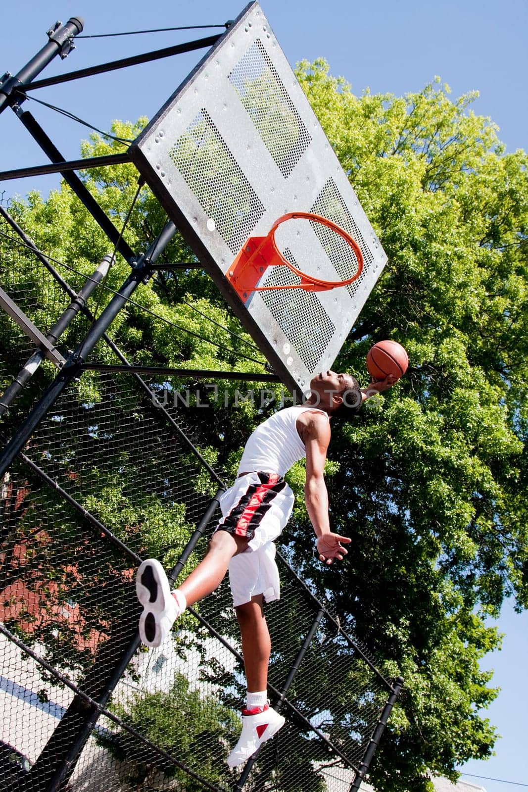 Sporty handsome African-American man dressed in white jumping in the air reaching for the basket while playing basketball in an outdoor court on a summer day trying to dunk the ball