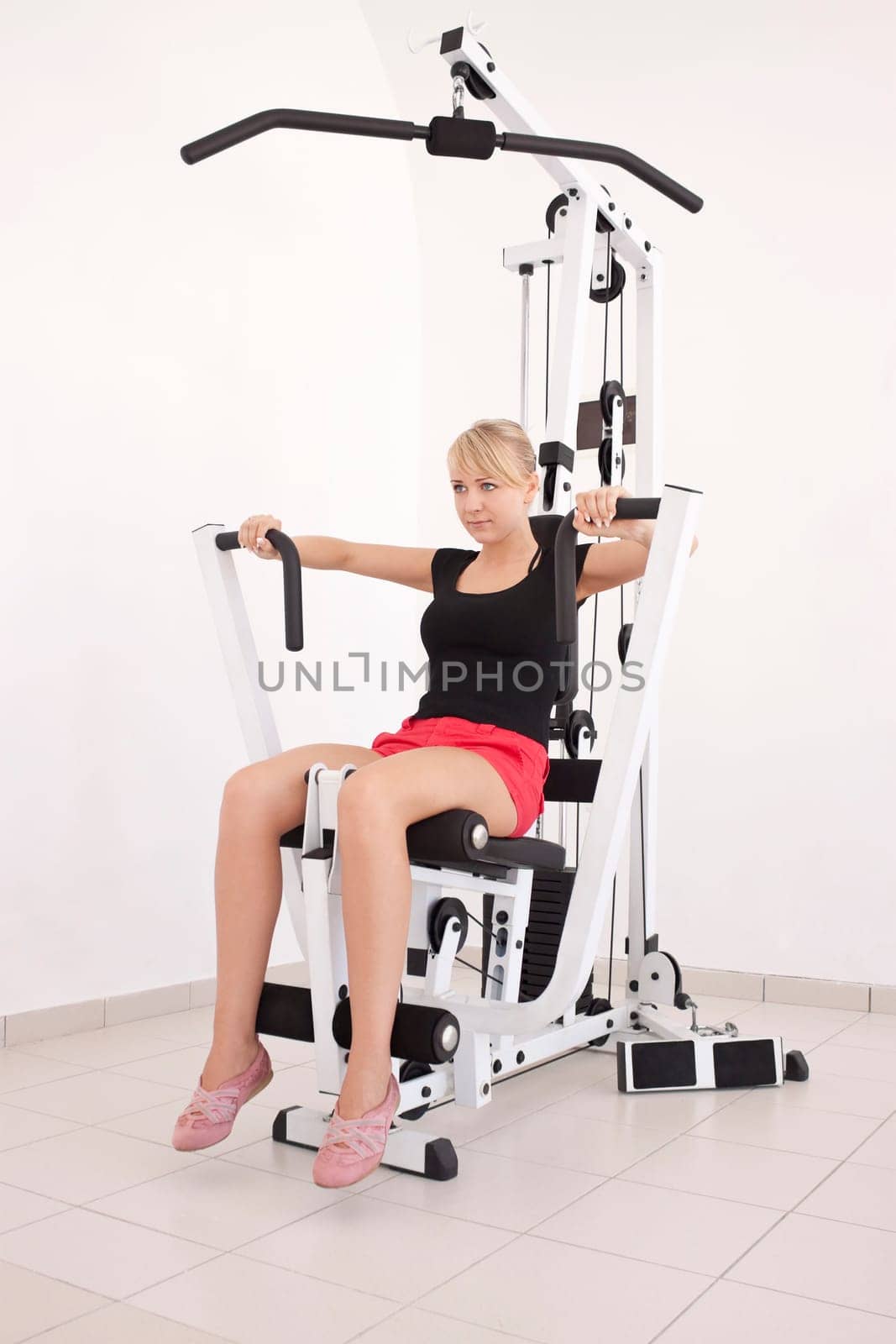 Young blond caucasian woman working out in gym. Pumping iron, lifting weight, pushing exercise. 3/4 view. Serious facial expression. Model looks aside.