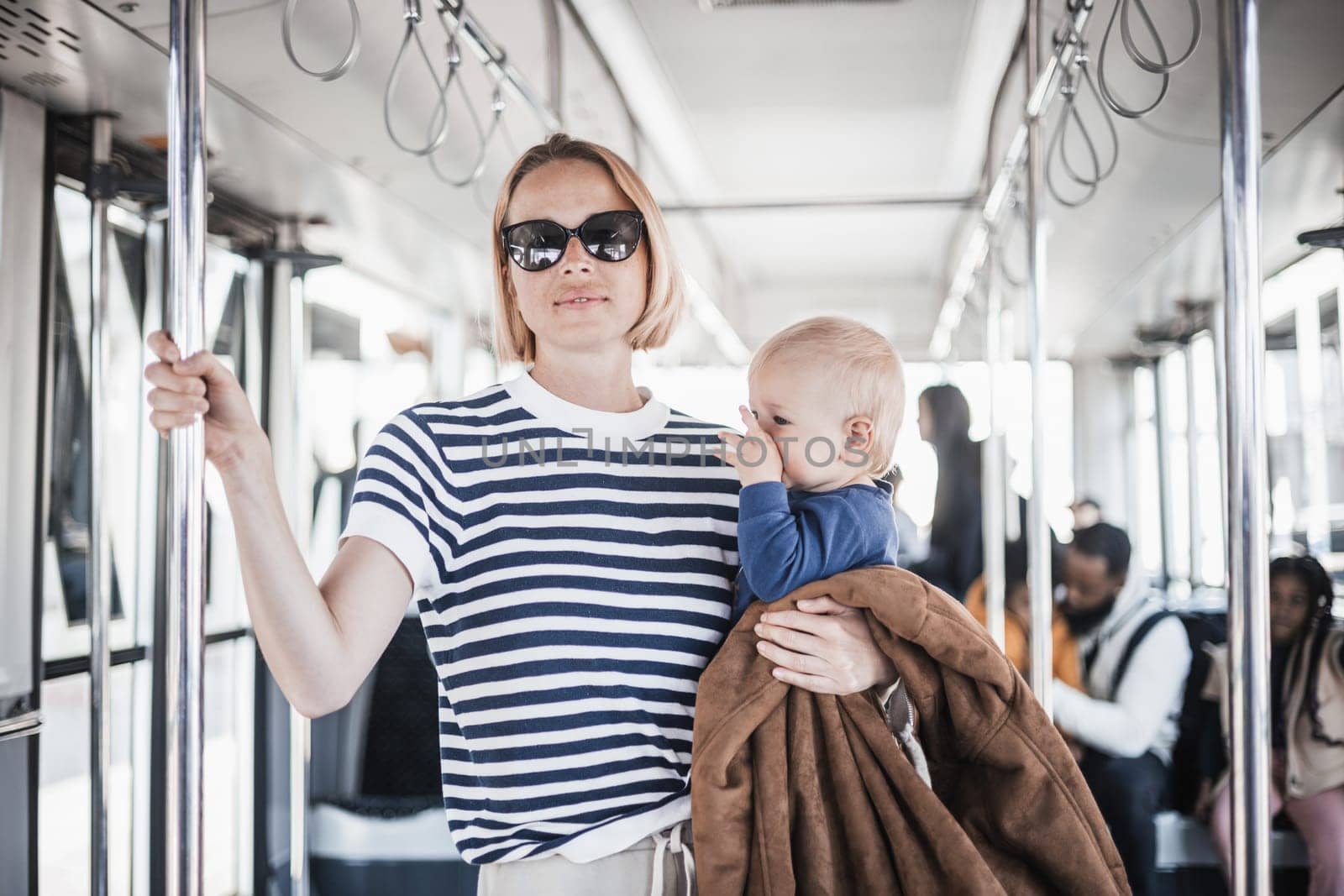 Mother carries her child while standing and holding on to bar holder on bus. Mom holding infant baby boy in her arms while riding in public transportation. Cute toddler traveling with mother