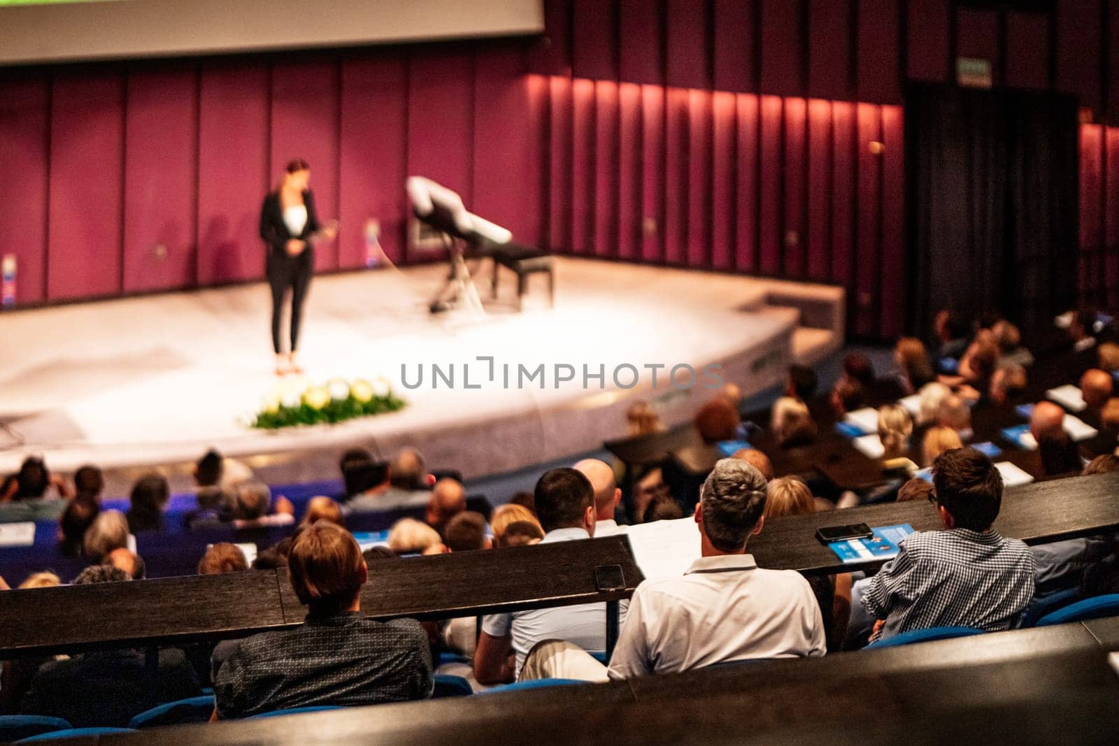 Business and entrepreneurship symposium. Female speaker giving a talk at business meeting. Audience in conference hall. Rear view of unrecognized participant in audience.