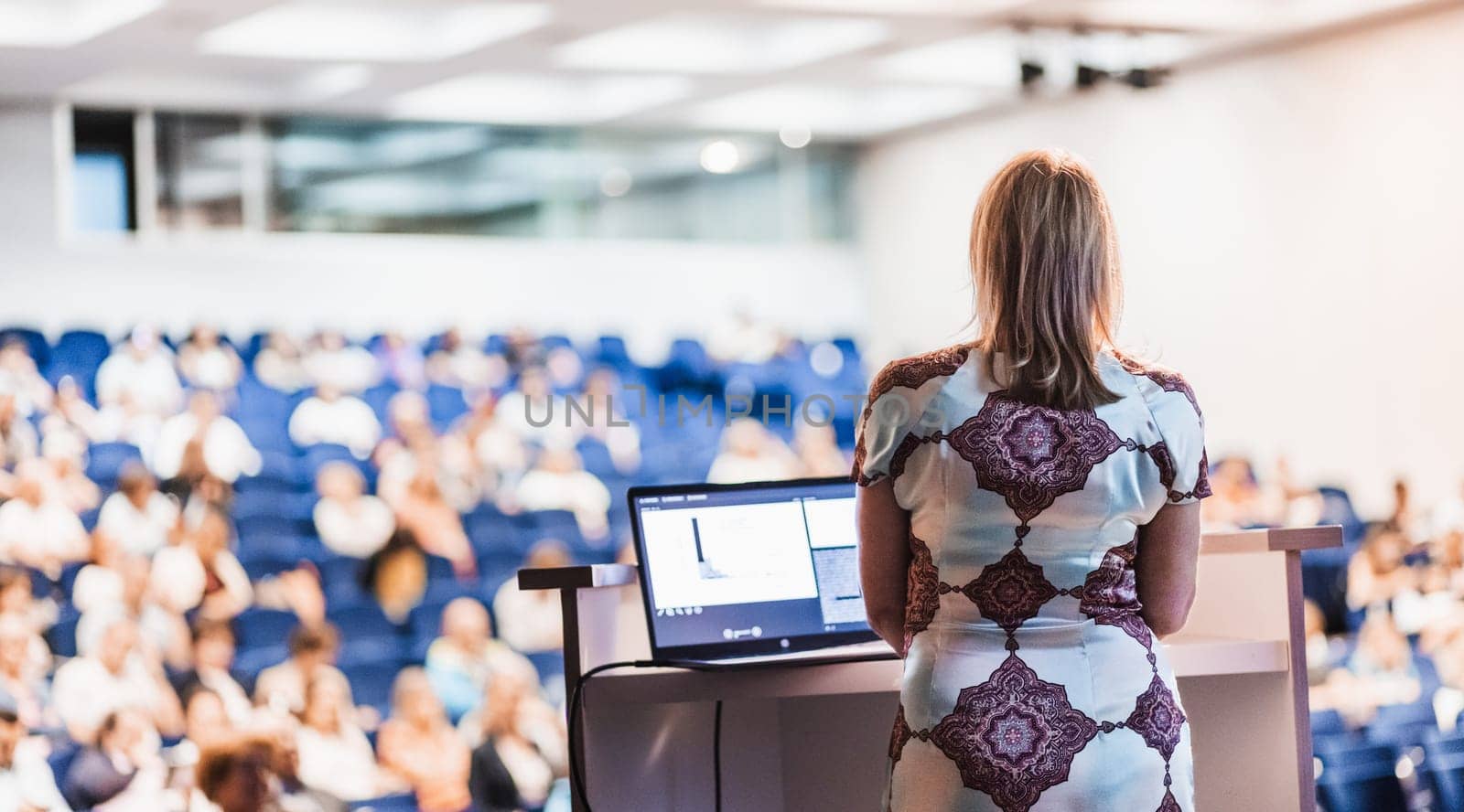 Female speaker giving a talk on corporate business conference. Unrecognizable people in audience at conference hall. Business and Entrepreneurship event.