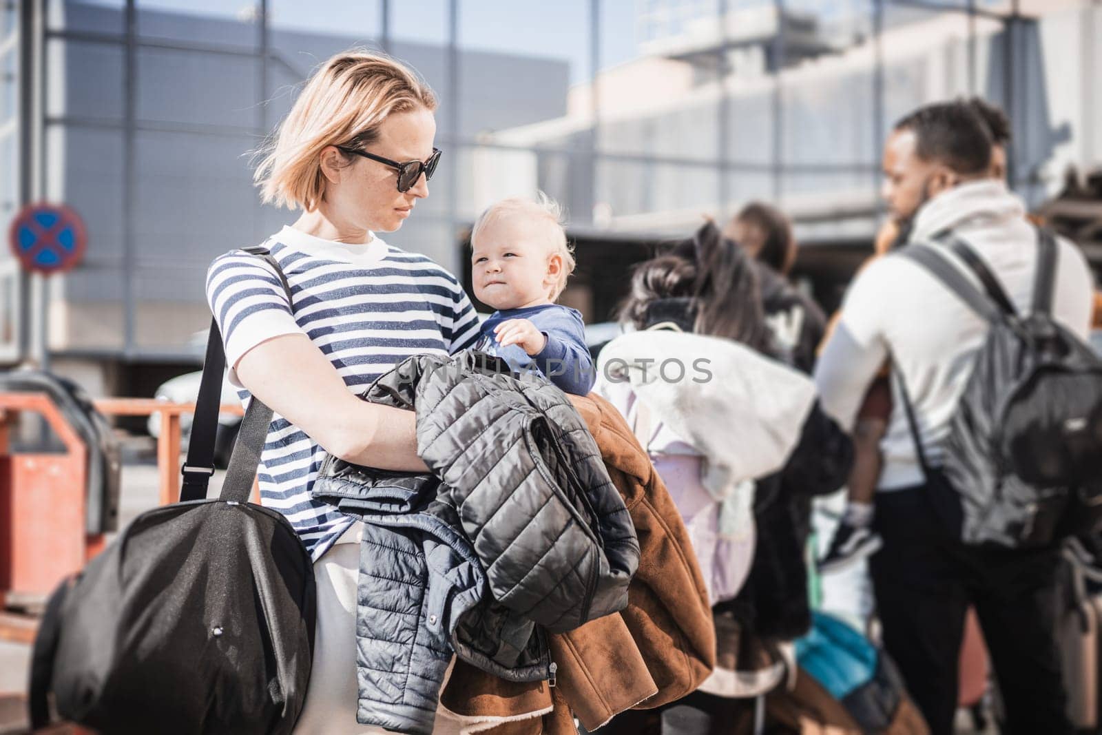 Motherat travelling with his infant baby boy child. Mom holding travel bag and her infant baby boy child while queuing for bus in front of airport terminal station.