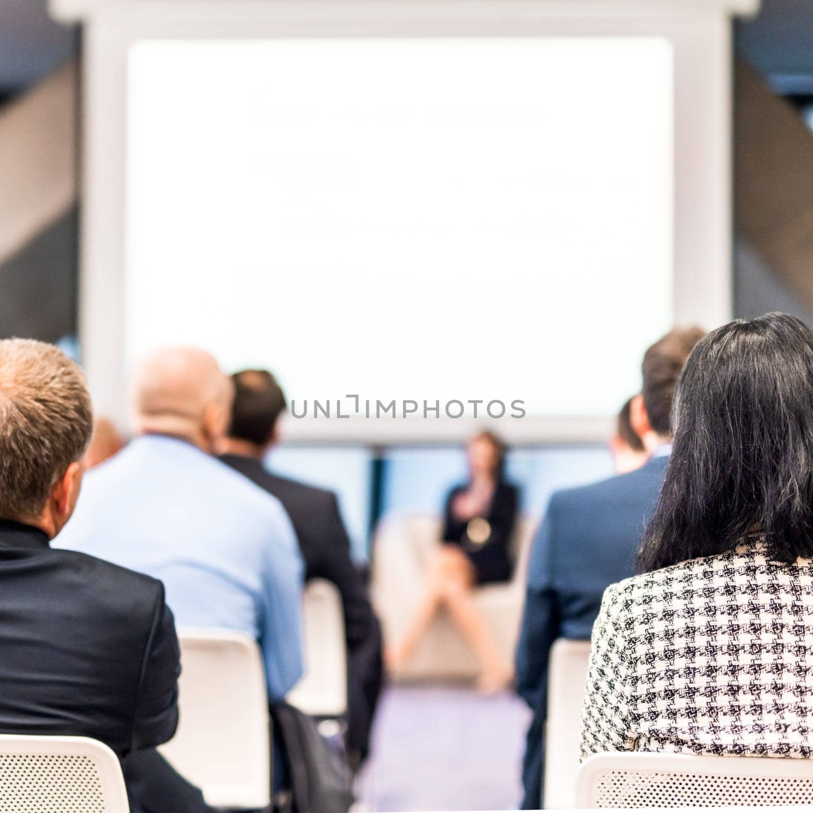 Business and entrepreneurship symposium. Female speaker giving a talk at business meeting. Audience in conference hall. Rear view of unrecognized participant in audience.