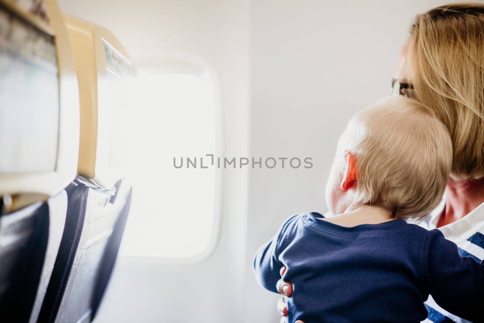 Mom and child flying by plane. Mother holding and playing with her infant baby boy child in her lap during economy comercial flight. Concept photo of air travel with baby. Real people