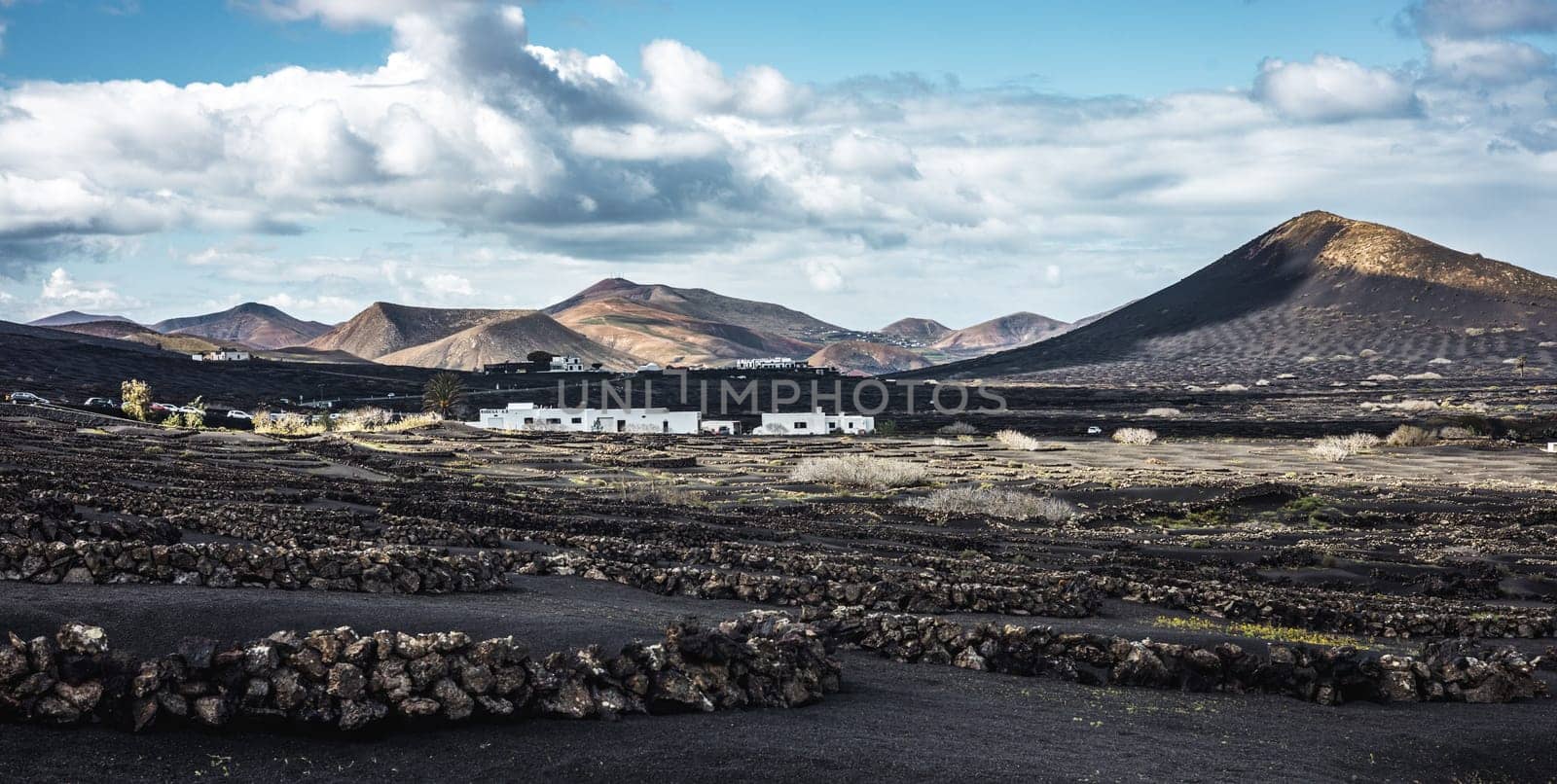 Traditional white houses in black volcanic landscape of La Geria wine growing region with view of Timanfaya National Park in Lanzarote. Touristic attraction in Lanzarote island, Canary Islands, Spain