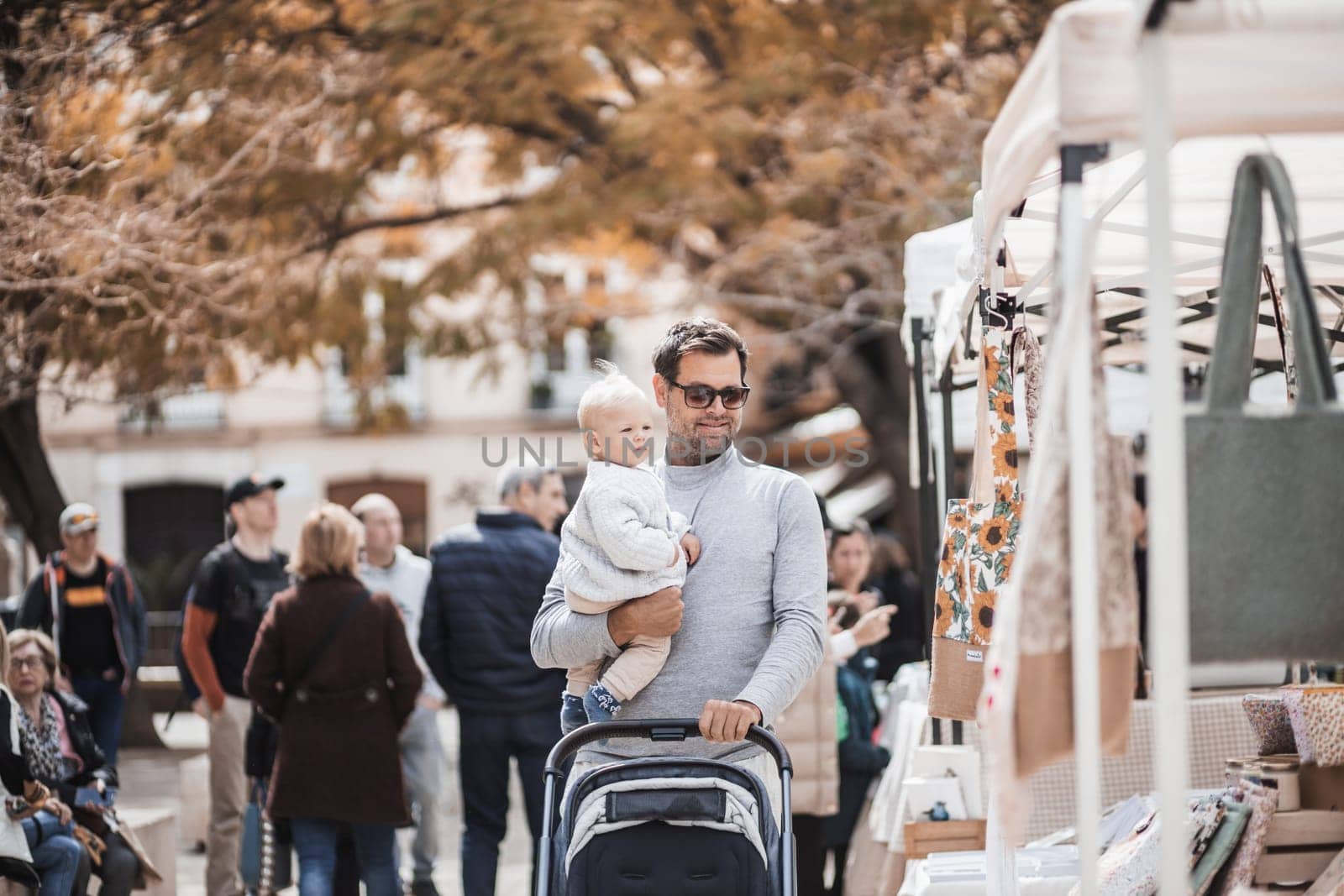 Father walking carrying his infant baby boy child and pushing stroller in crowd of people wisiting sunday flea market in Malaga, Spain