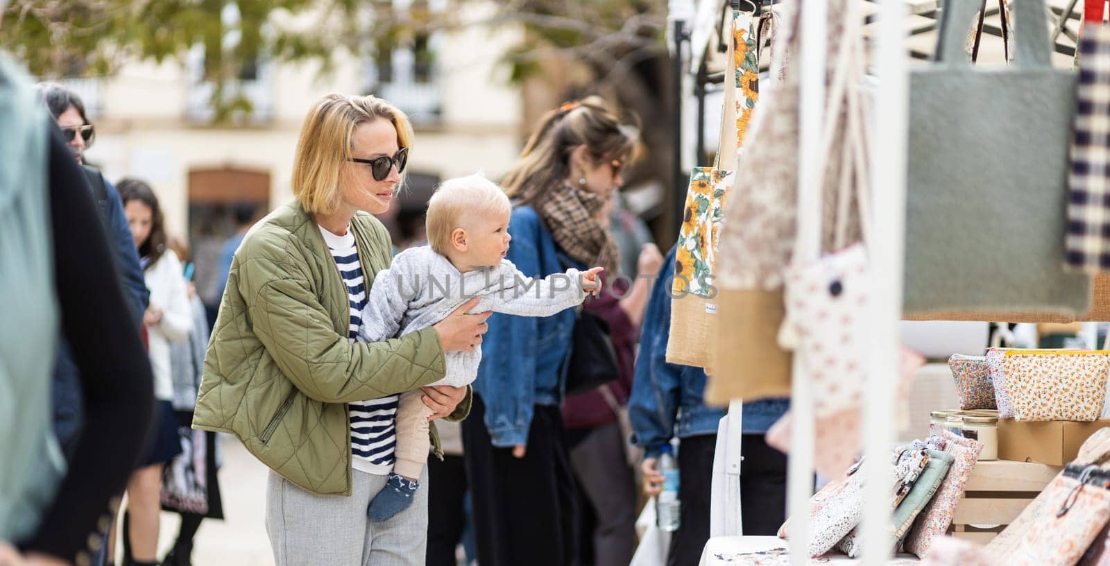 Mother walking carrying his infant baby boy child in crowd of people wisiting sunday flea market in Malaga, Spain.