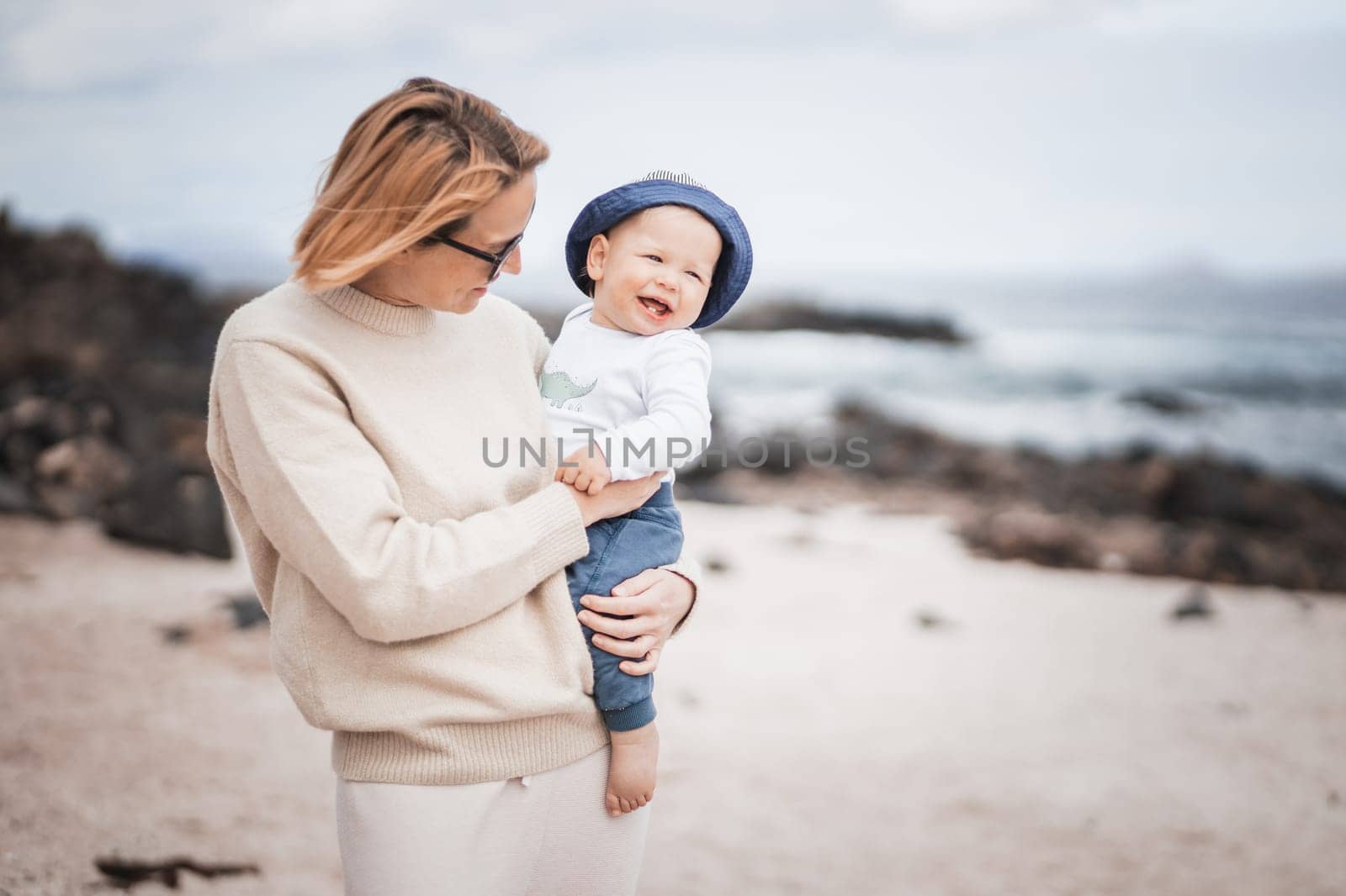 Mother enjoying winter vacations holding and playing with his infant baby boy son on volcanic sandy beach on Lanzarote island, Spain. Family travel and vacations concept.