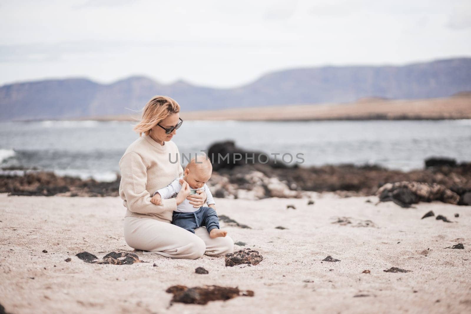 Mother enjoying winter beach vacations playing with his infant baby boy son on wild volcanic sandy beach on Lanzarote island, Canary Islands, Spain. Family travel and vacations concept.
