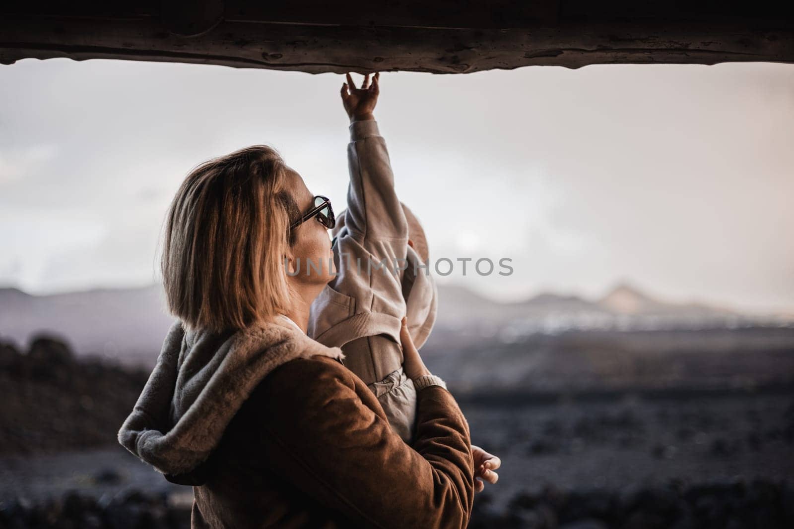 Mother enjoying winter vacations playing with his infant baby boy son on black sandy volcanic beach of Janubio on Lanzarote island, Spain on windy overcast day. Family travel vacations concept