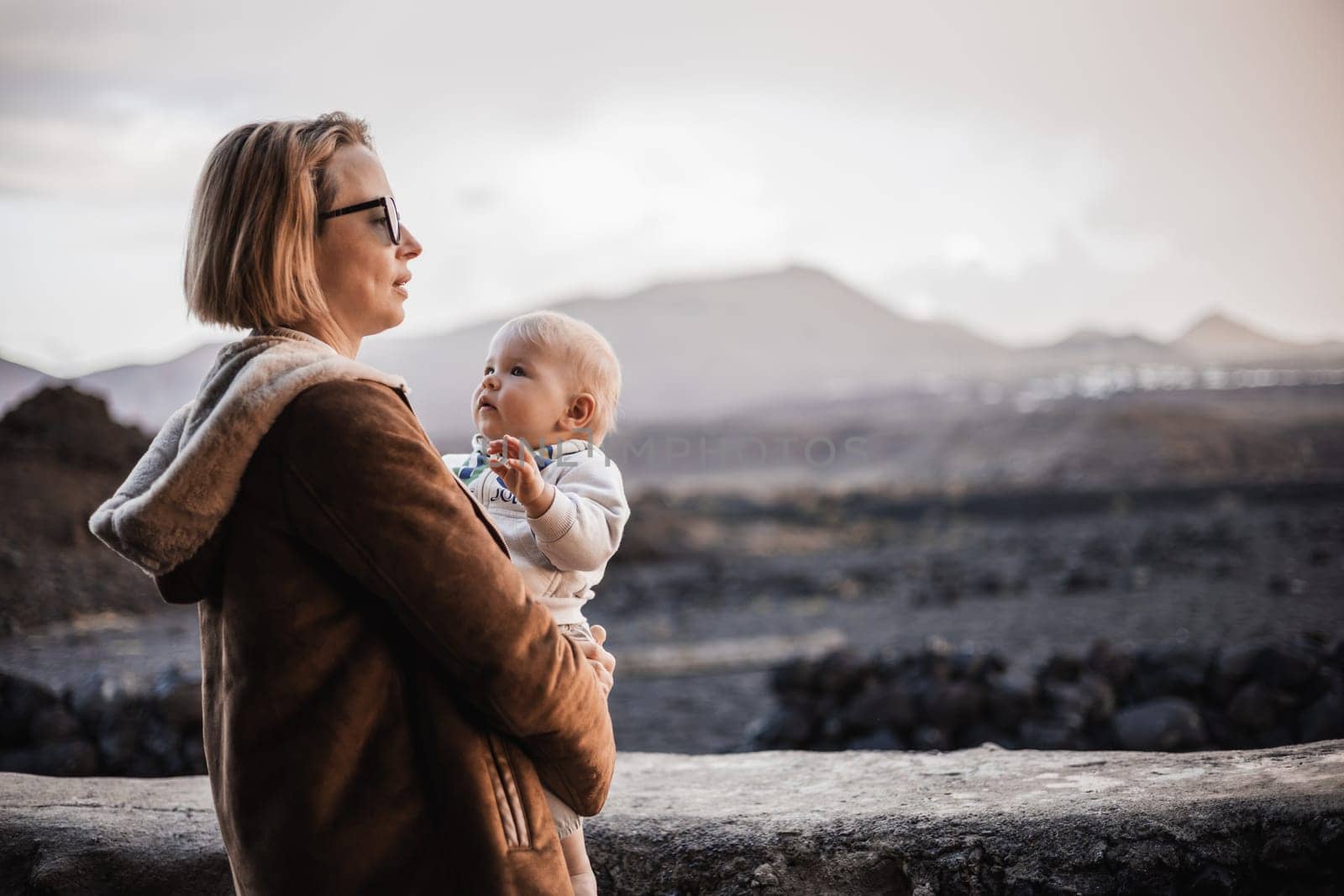 Mother enjoying winter vacations playing with his infant baby boy son on black sandy volcanic beach of Janubio on Lanzarote island, Spain on windy overcast day. Family travel vacations concept