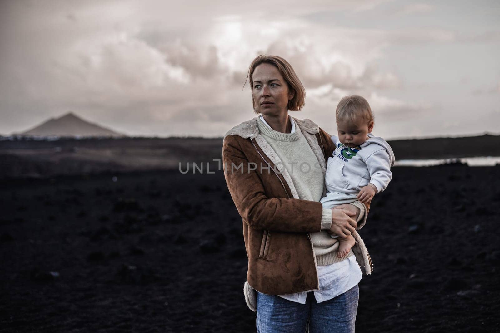 Mother enjoying winter vacations playing with his infant baby boy son on black sandy volcanic beach of Janubio on Lanzarote island, Spain on windy overcast day. Family travel vacations concept
