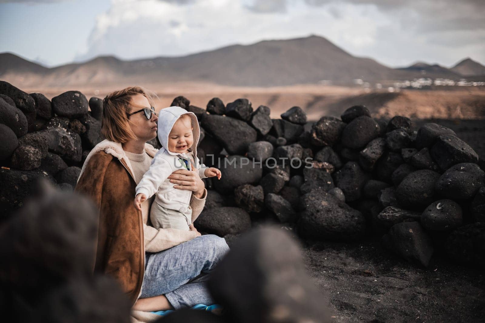 Mother enjoying winter vacations playing with his infant baby boy son on black sandy volcanic beach of Janubio on Lanzarote island, Spain on windy overcast day. Family travel vacations concept