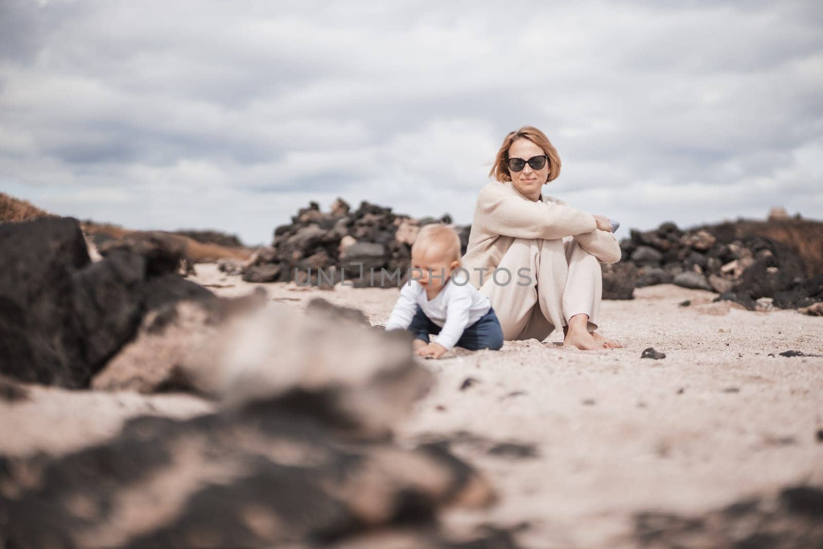 Mother enjoying winter beach vacations playing with his infant baby boy son on wild volcanic sandy beach on Lanzarote island, Canary Islands, Spain. Family travel and vacations concept.