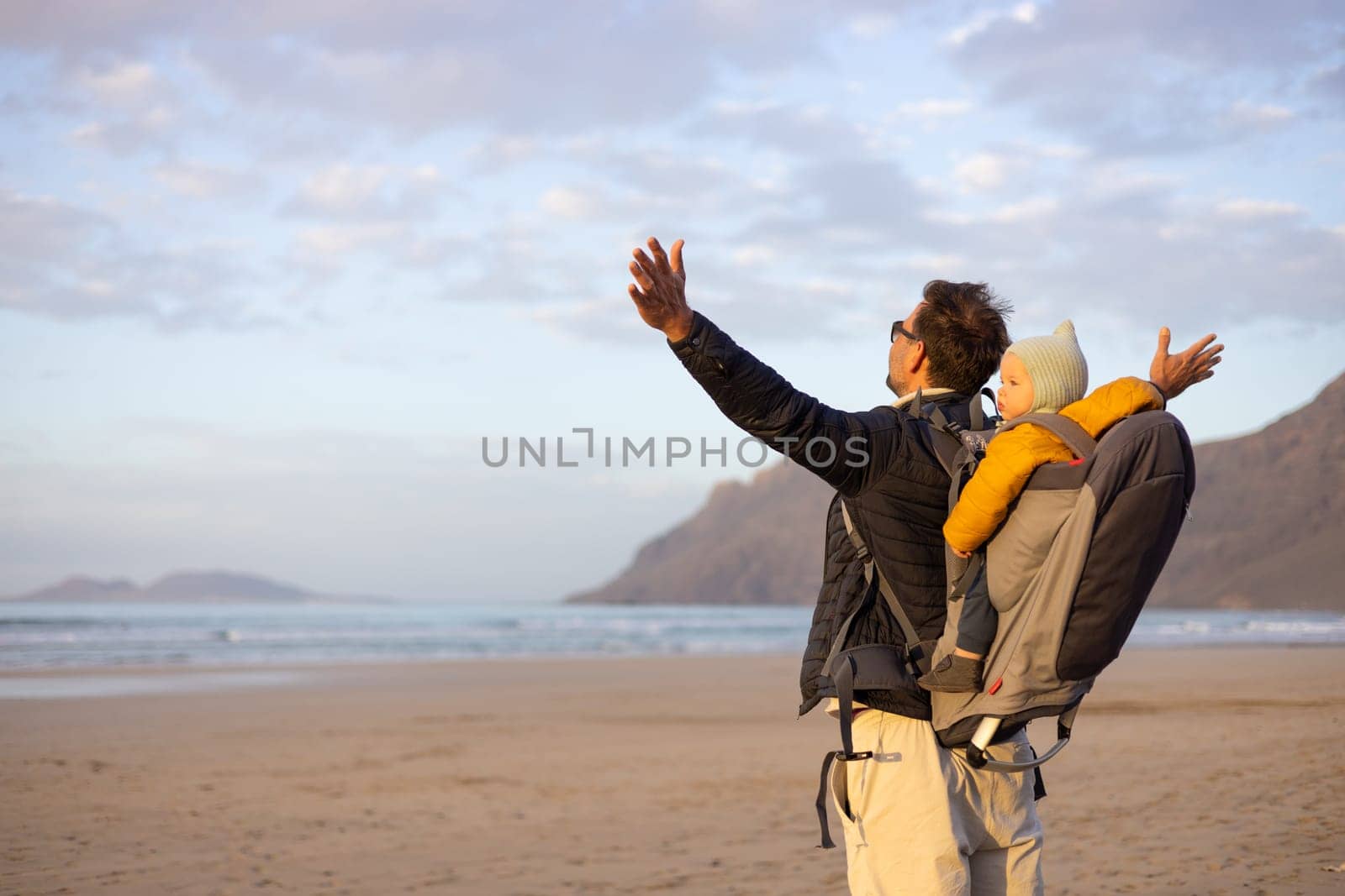 Father rising hands to the sky while enjoying pure nature carrying his infant baby boy son in backpack on windy sandy beach of Famara, Lanzarote island, Spain at sunset. Family travel concept