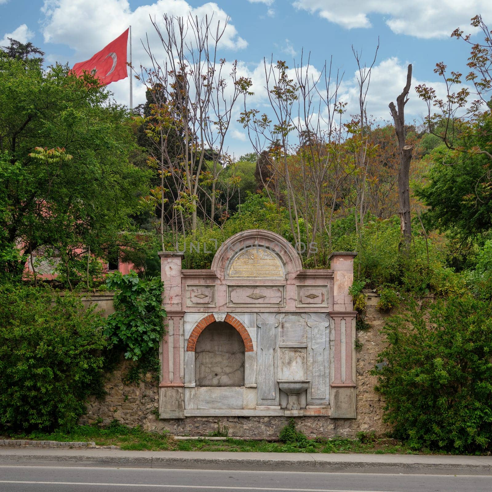 Kadin Efendi Cesmesi, a beautiful fountain house in Besiktas, Istanbul, Turkey, made of carved marble and stone, surrounded by dense green trees, with Turkish flag in the background