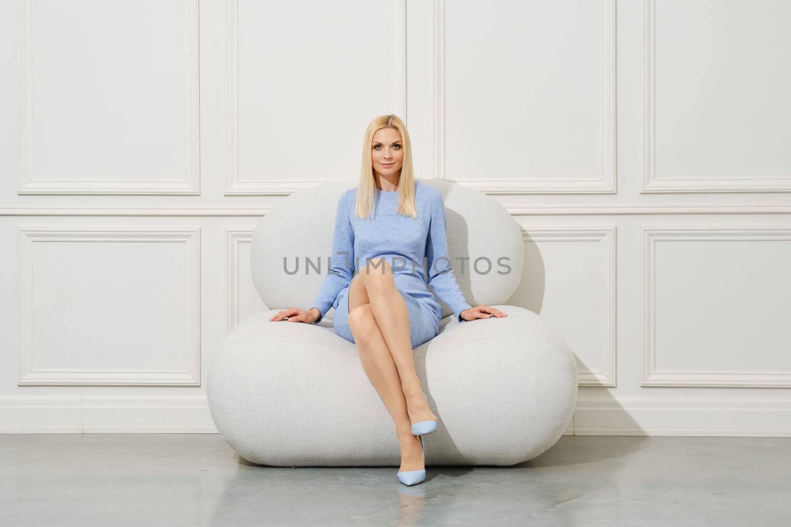 Pretty woman in light blue tight knit dress sitting on soft armchair with crossed legs