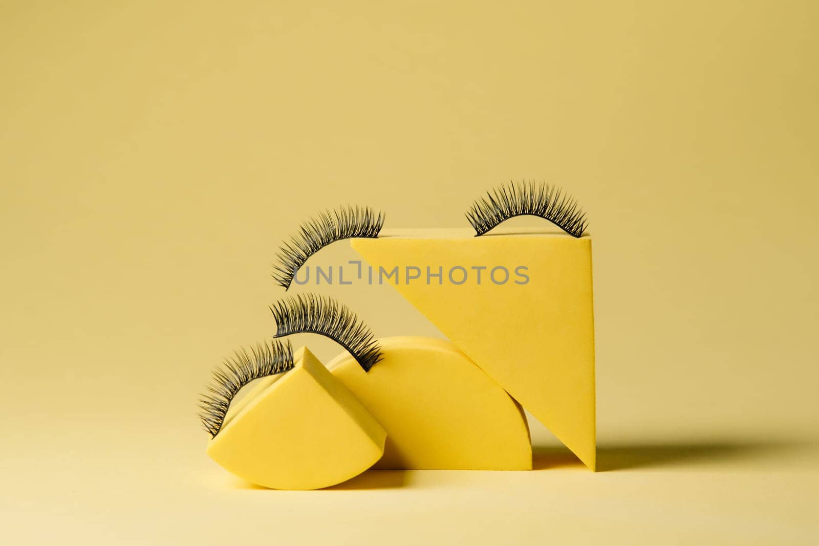Fashionable black artificial eyelashes sticking on tops of various bright yellow shapes against xanthous background. Fake lashes for creating stylish image in process of cilia extension.