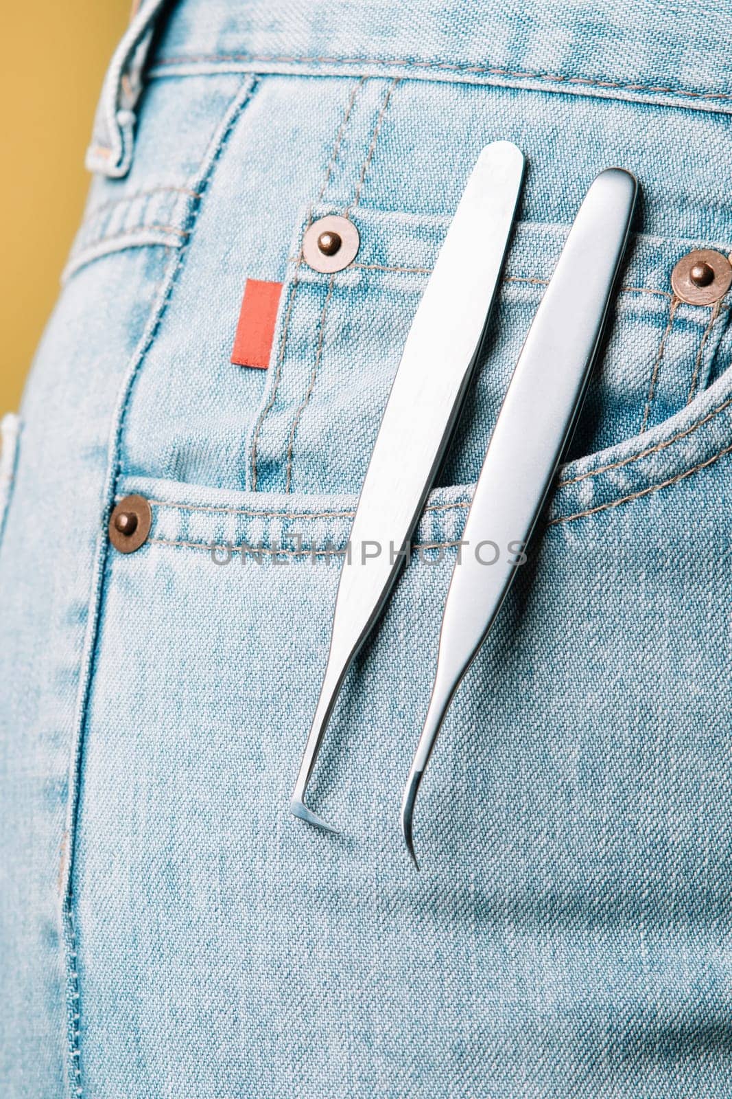 Pair of high-quality tweezers, using for isolating and gluing lashes, assisting to create charming look, hanging from pocket of blue denim jeans of unrecognizable woman. Vertical.