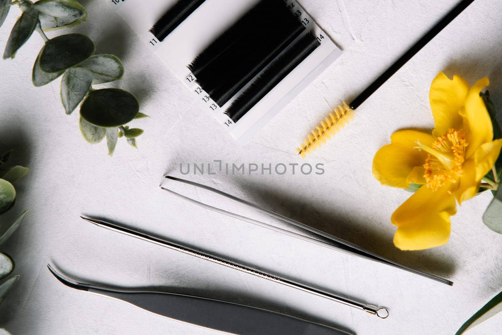 View from above of different professional tools using for fake eyelash extension procedure, isolated tweezers, palette of cilia and brush, lying on white table near green stems and yellow flower.