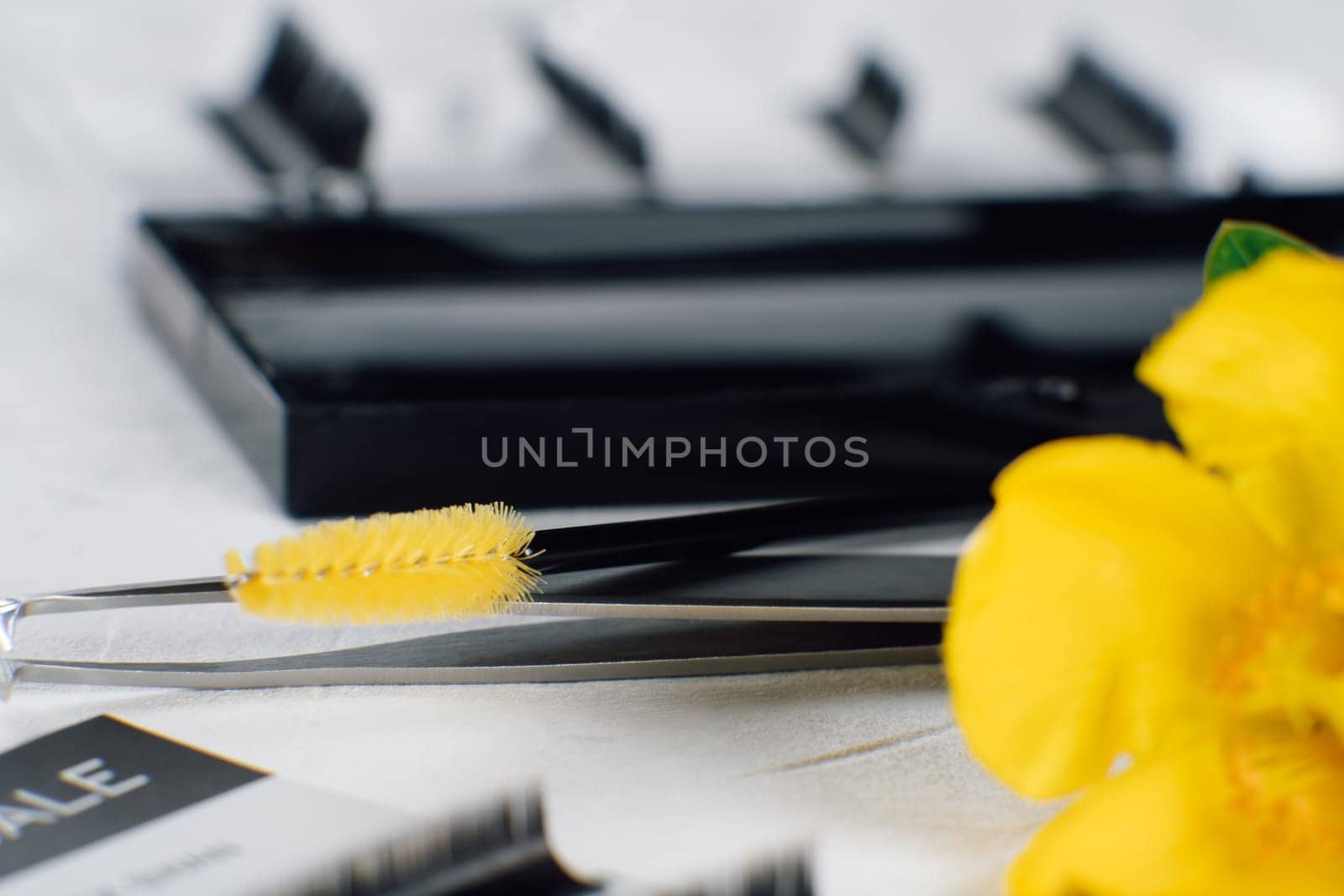 Bright flower, left on foreground, slightly covering disposable eyelash extension brush with yellow delicate bristles lying on isolated tweezers on white table full of tools for procedure.