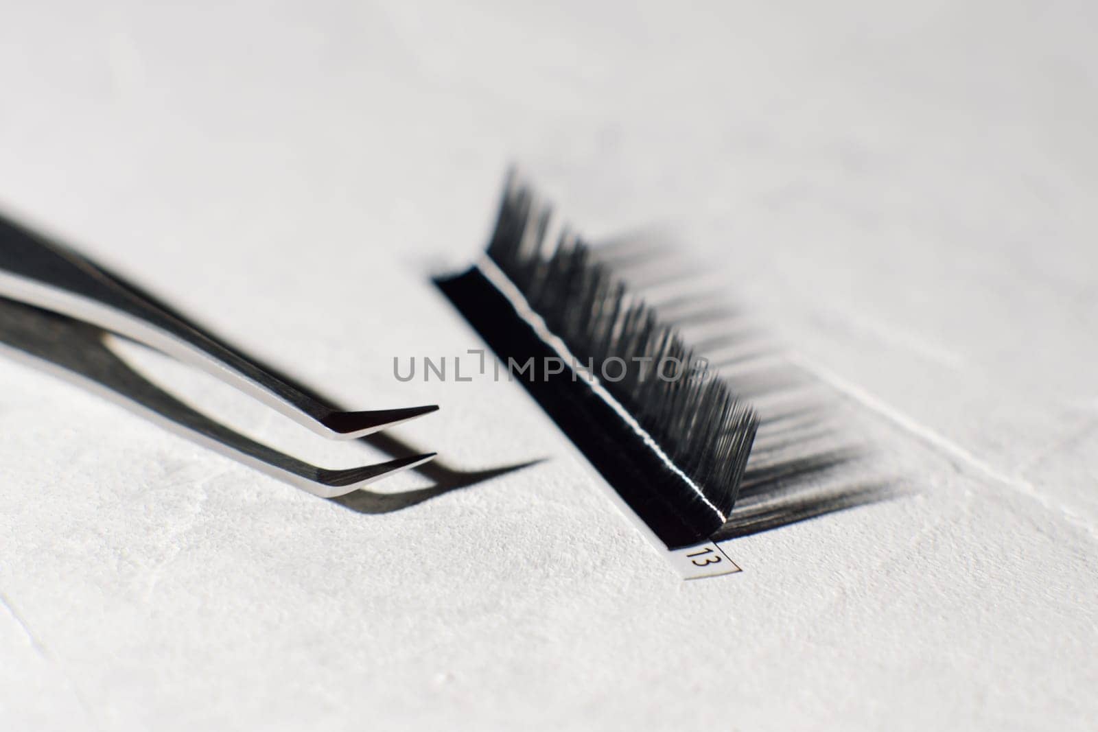 Tweezers with sharp tip, using for isolating small lashes, lying near palette of black artificial eyelashes on white table with cast shandows of tools. Instruments for cilia extension procedure.