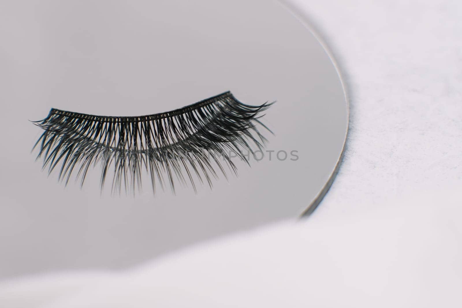 Black and long artificial eyelashes using for lash extension procedure lying on round mirror on white table. Isolated fake cilia reflecting in looking-glass, making image of fluffiness and volume.