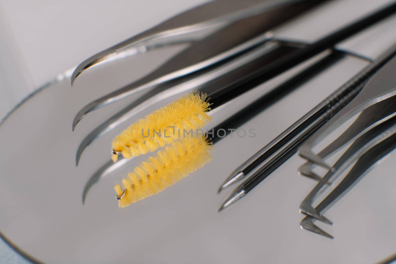 Various professional tools for eyelash extension procedure left on round mirror on table. Tweezers, yellow disposable brush and pen reflecting in looking-glass, lying on desk with nobody.