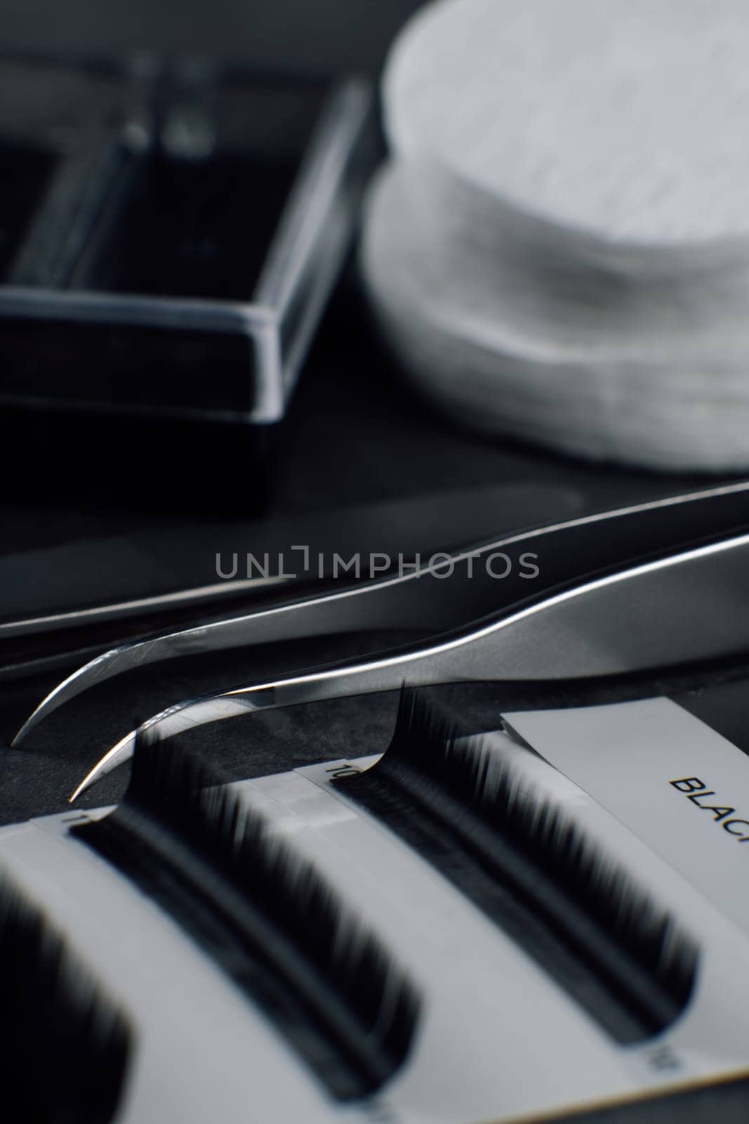 Close up of black artificial eyelashes in palette lying close to isolated tweezers on table prepared for lash extension procedure. Different tools and consumable for creating flawless look. Vertical.