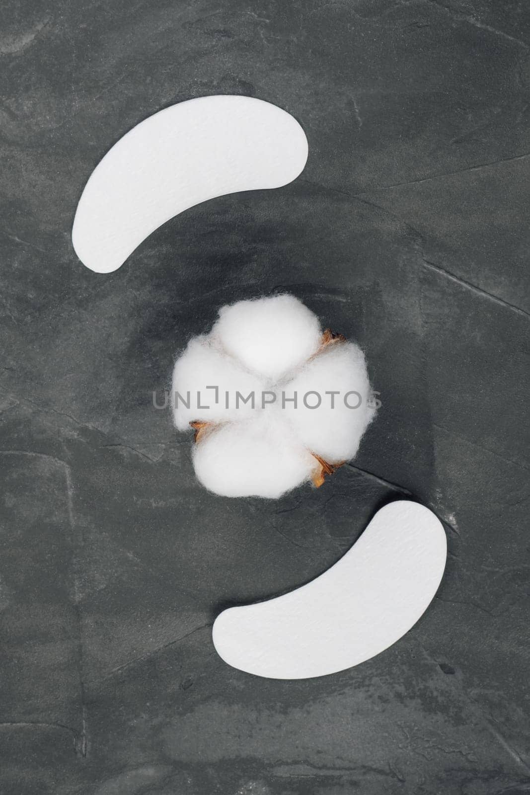 View from above of pair of white patches, applying on lower eyelids and isolating lashes, using for eyelash extension procedure lying out on gray table around cotton pads in form of flower. Vertical.