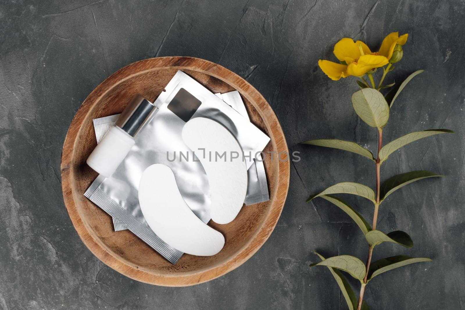 View from above of disposable patches for eyelash extension procedure lying in wooden bowl full of materials on gray table near yellow flower. White consumable for isolating lashes in round plate.