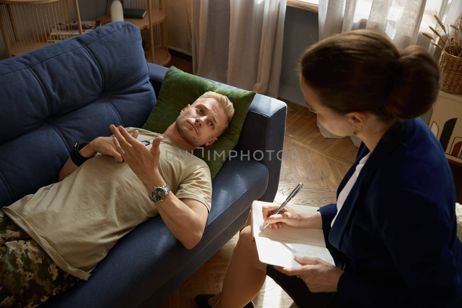 Frustrated sad soldier with ptsd talking at psychiatrist and gesturing while lying on couch during therapy session. Overhead view