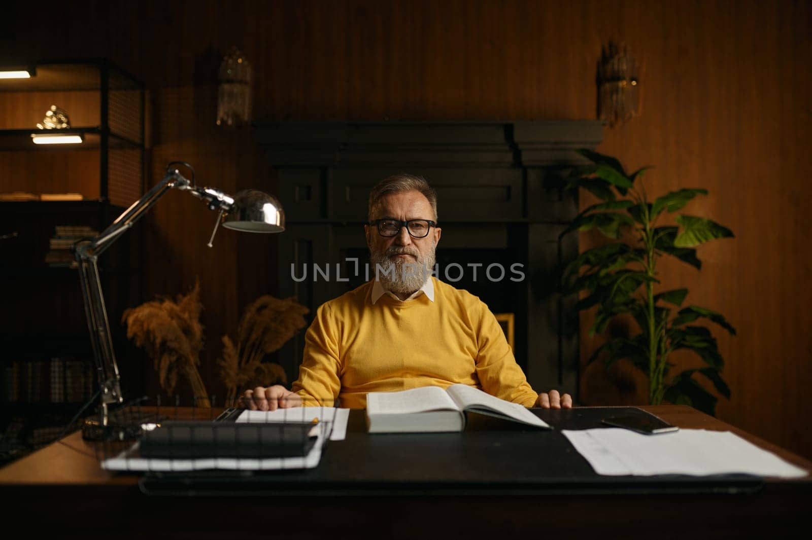 Portrait of serious pensive senior man on retirement working at home office in evening. Elderly male sitting at table taking break while reading book at library
