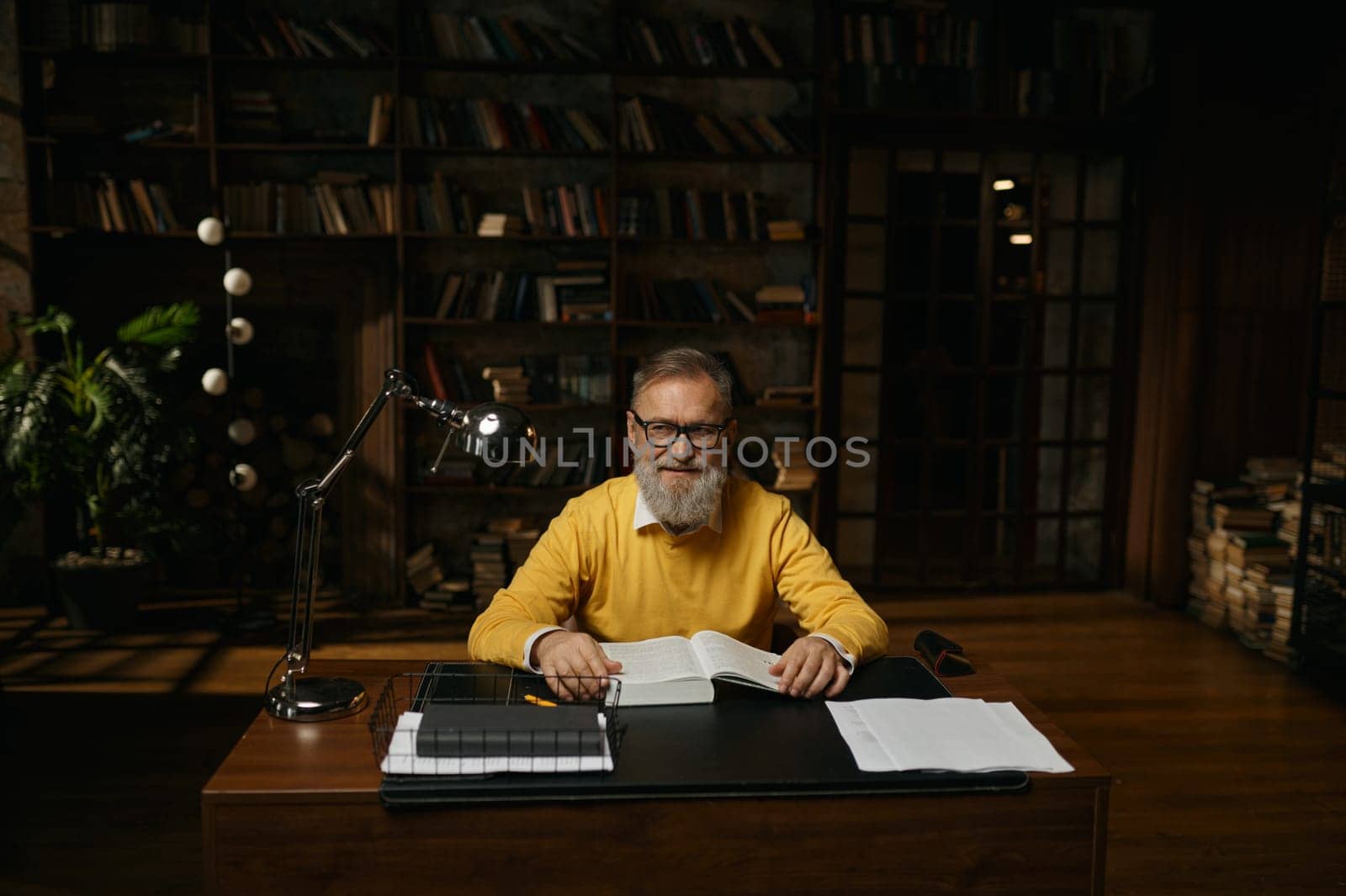 Senior man working with documents at home office. Elderly male sitting at table in library looking at camera