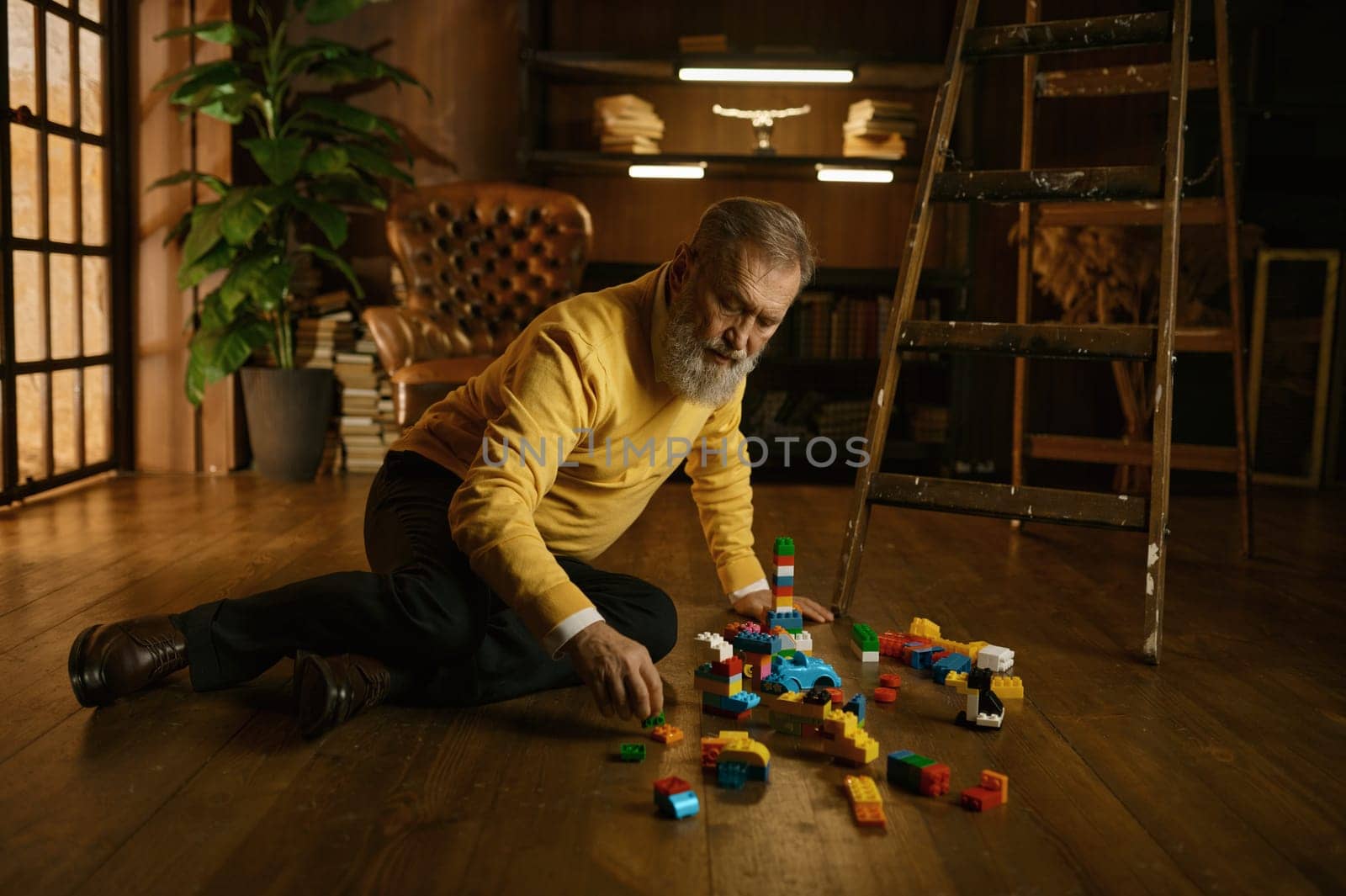 Senior old man playing toy building blocks while sitting on home floor during cozy evening. Handsome adult businessman on retirement