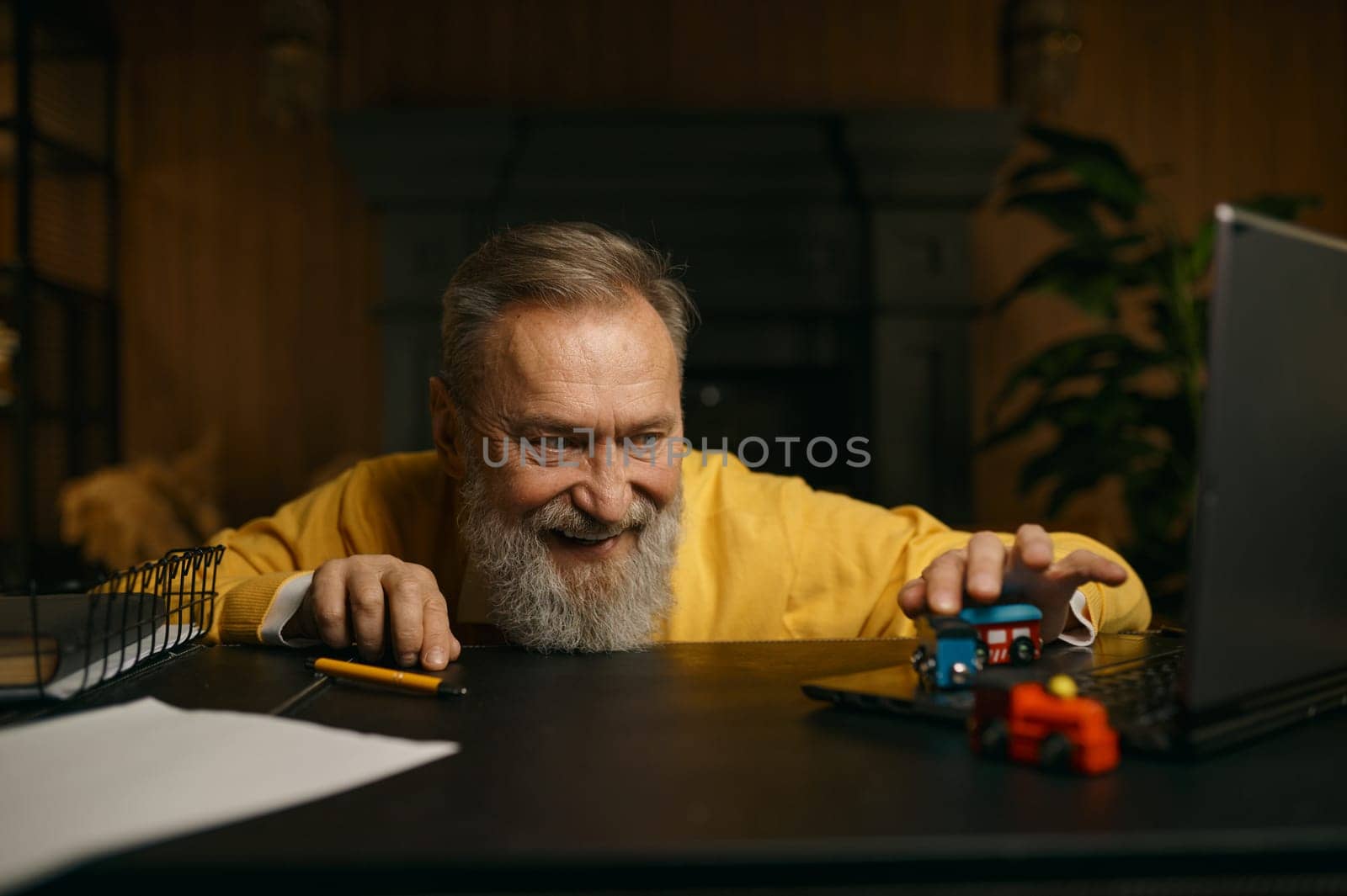 Selective focus portrait of senior man playing with toy train at workplace with laptop computer on desk. Mature older male dreaming about vacation time and railway trip on weekend